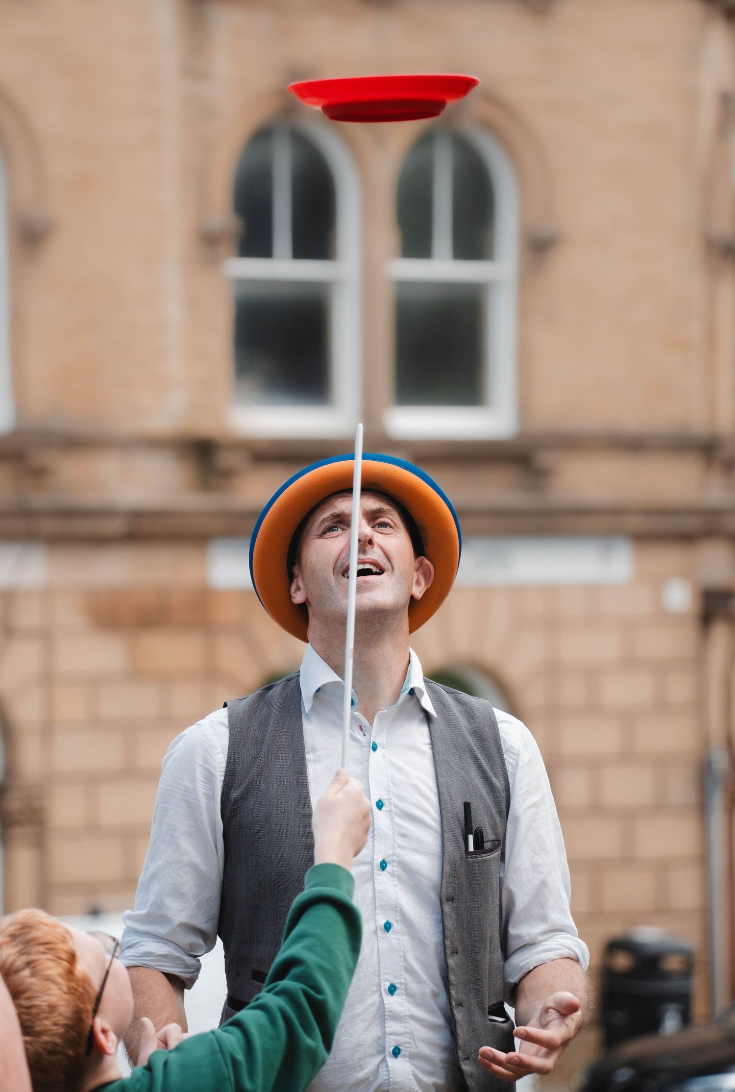 A street performer balances a spinning red plate on a stick while smiling outdoors.