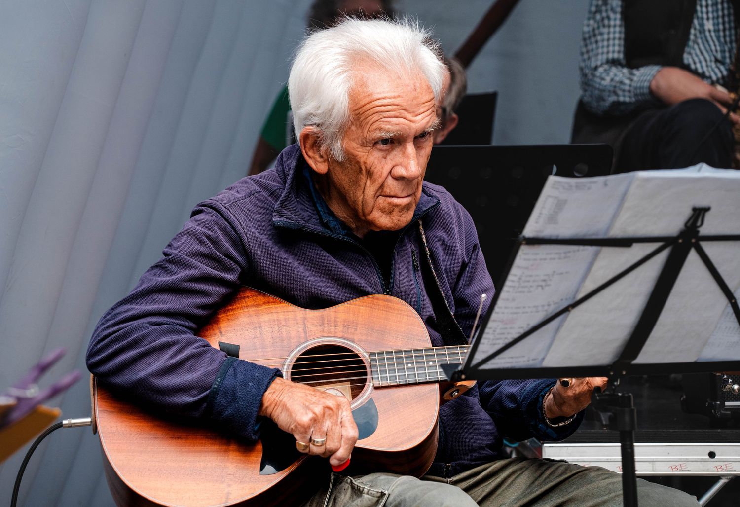 Elderly man with white hair playing acoustic guitar at outdoor music performance.