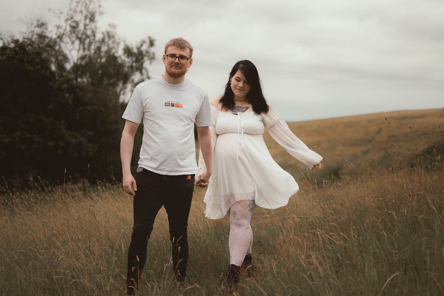 Couple holding hands in golden meadow, pregnant woman in white dress with man in white t-shirt.