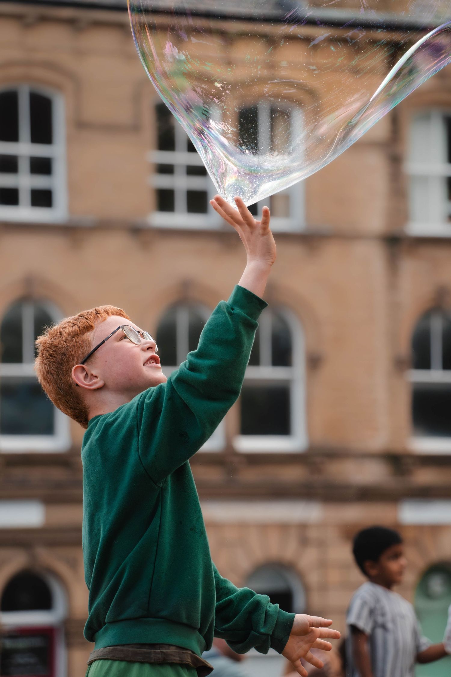 A red-haired boy wearing glasses reaches up to touch a giant soap bubble outdoors.