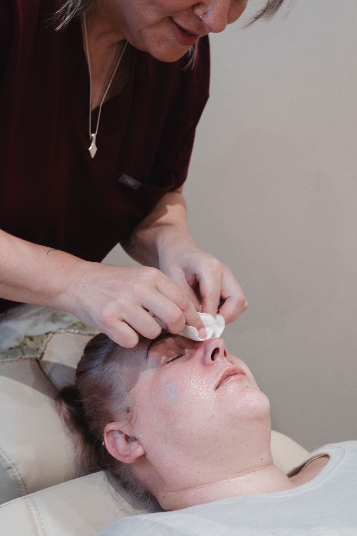 Beauty therapist in burgundy scrubs performing facial treatment on reclined client.