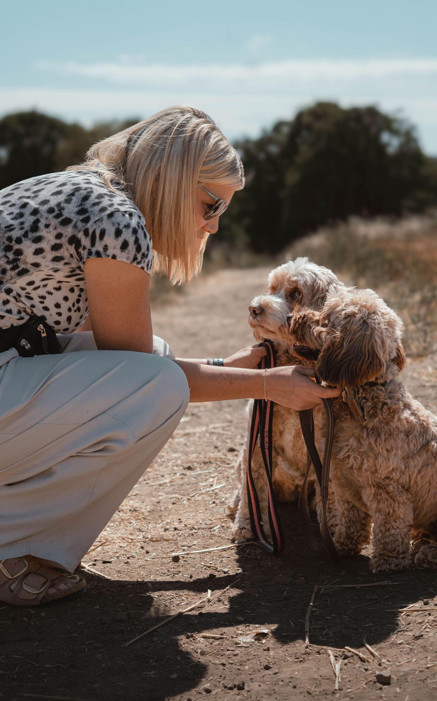Blonde woman in spotted top crouching and petting two small Cockapoo dogs on a dirt path.