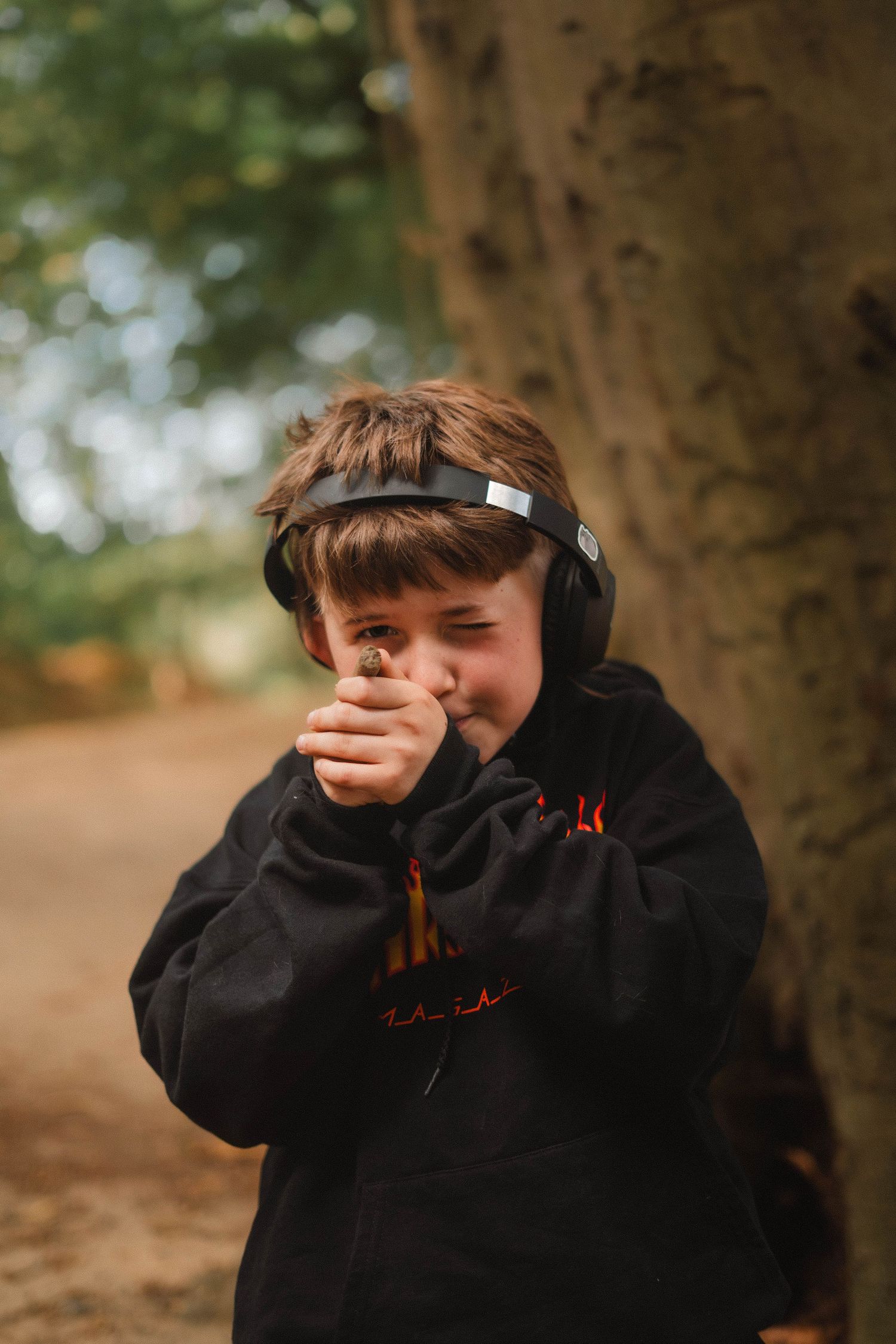 A young boy wearing headphones covers his mouth while standing near a tree outdoors.