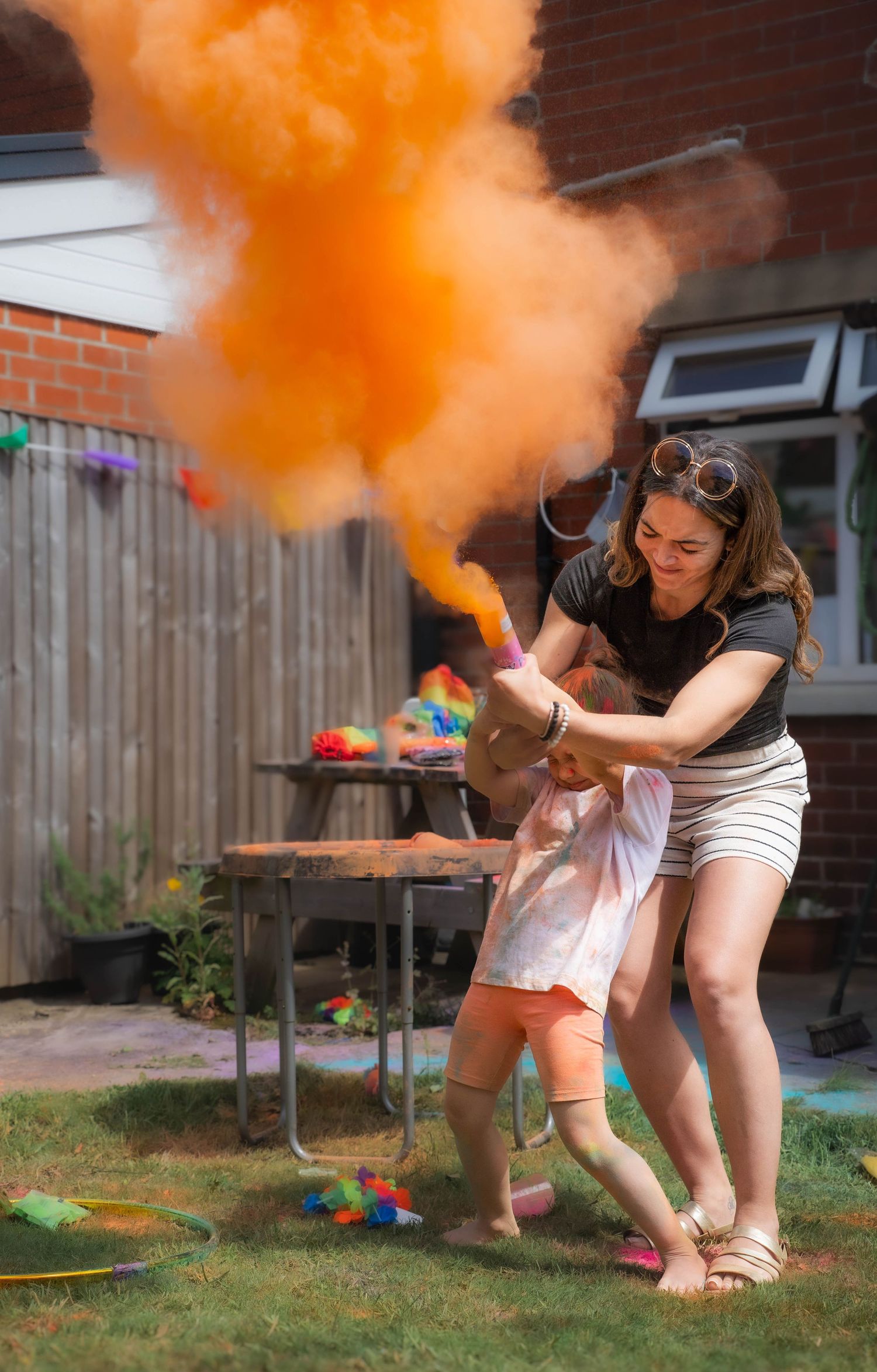 Woman and young child releasing a large cloud of orange Holi powder in a sunny backyard.