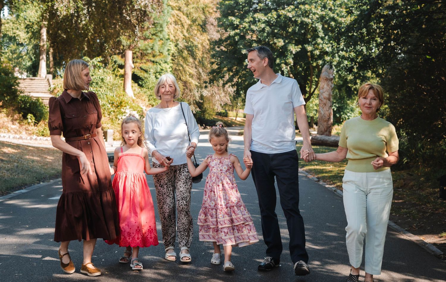 A multi-generational family walks together along a sunny tree-lined path outdoors.