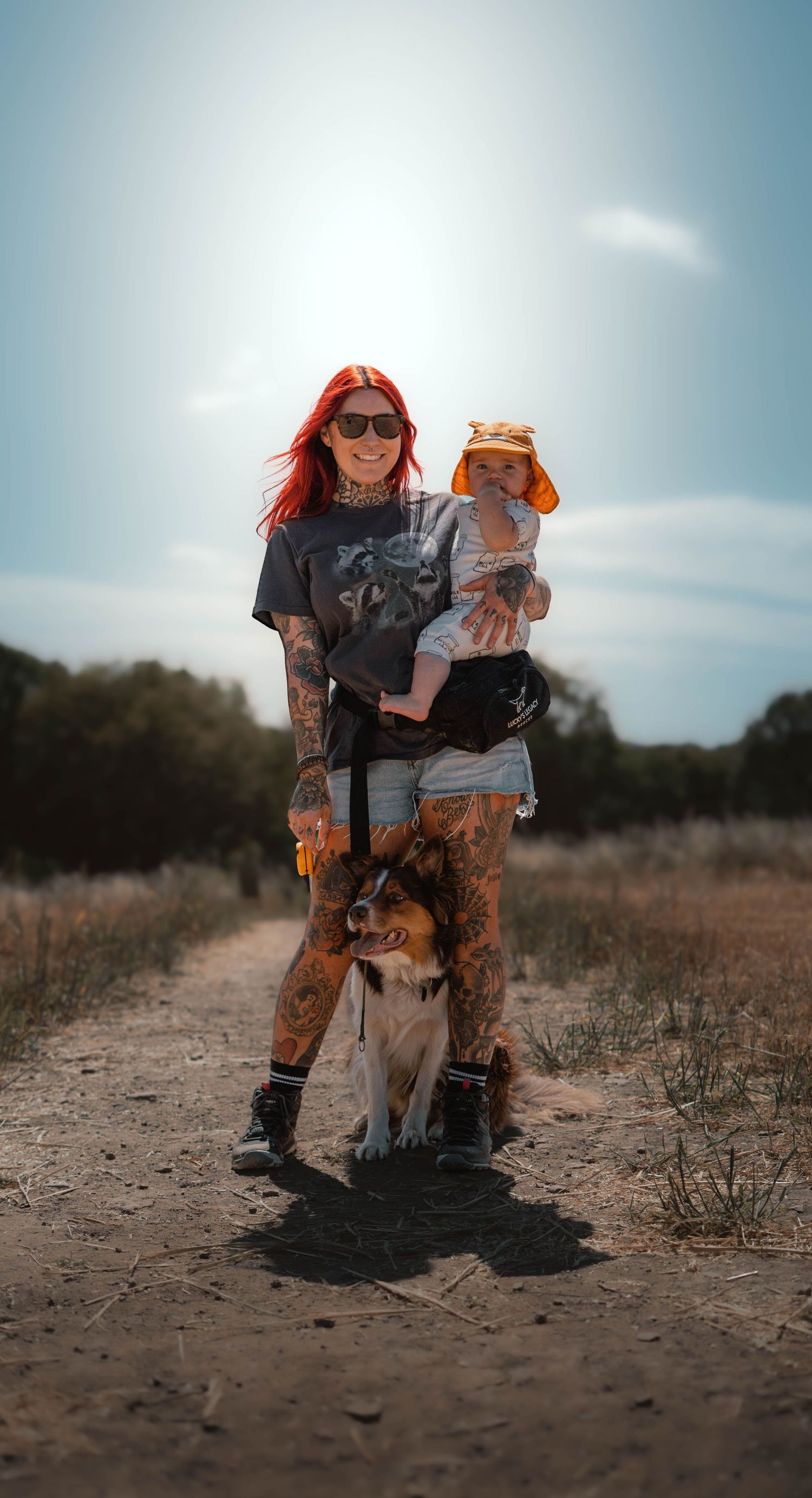 Tattooed woman with red hair holds baby while standing with a collie dog on a sunny dirt trail.