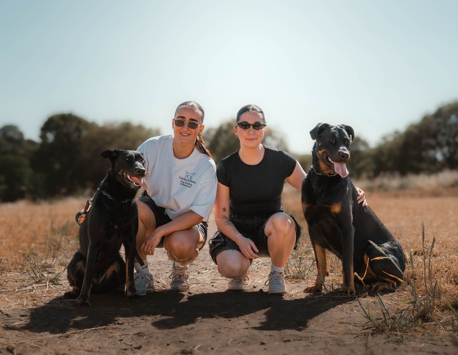 Two women crouch with two black Doberman dogs on a sunny dirt field with sparse trees in the background.