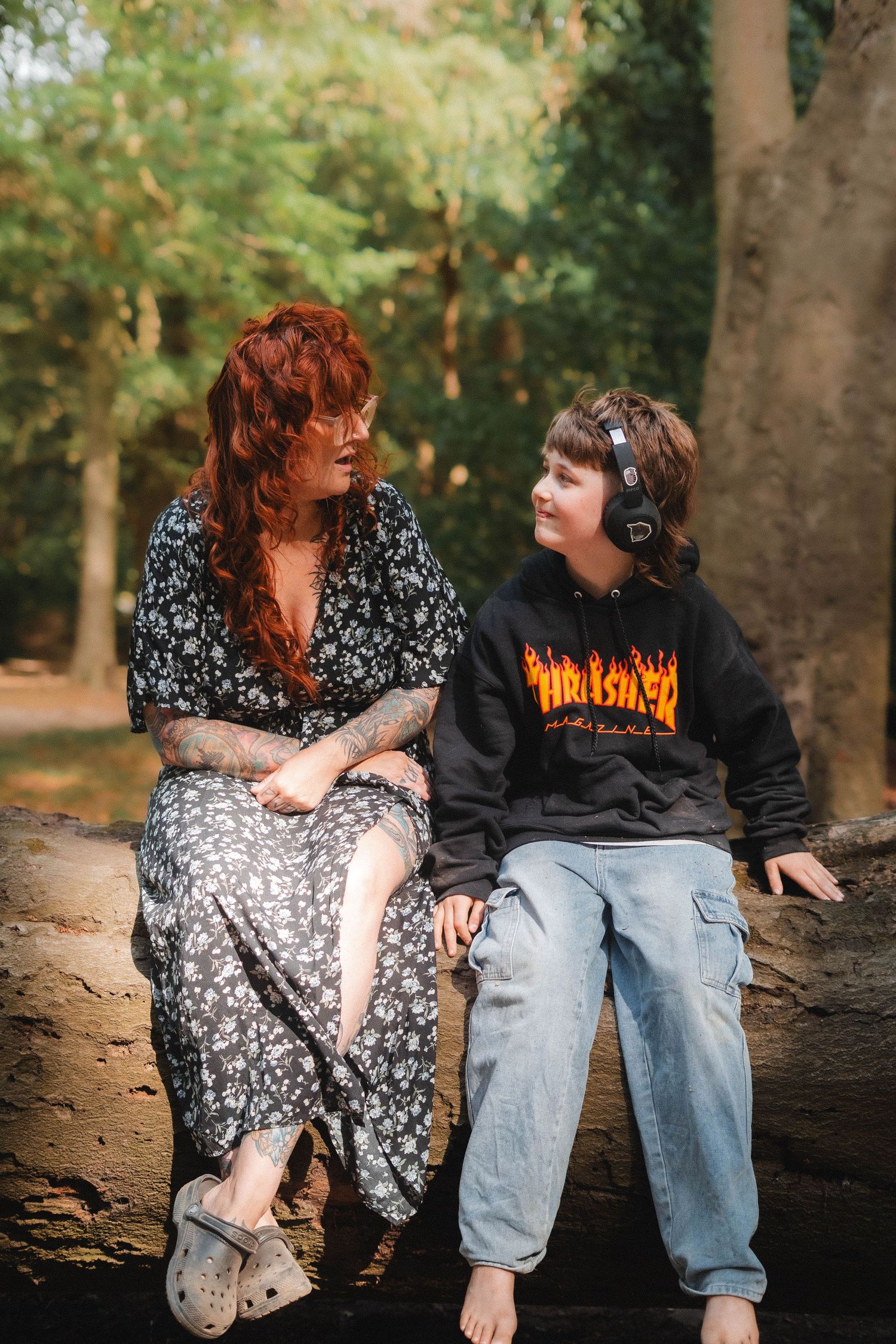 A woman and boy sit on a log in a forest, the boy wearing a Thrasher hoodie and headphones.