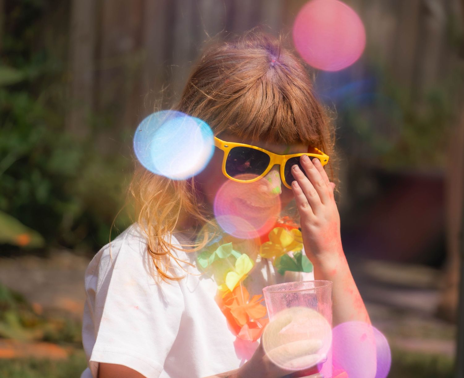 Young girl wearing yellow sunglasses and floral lei holds a drink outdoors in sunny backyard.