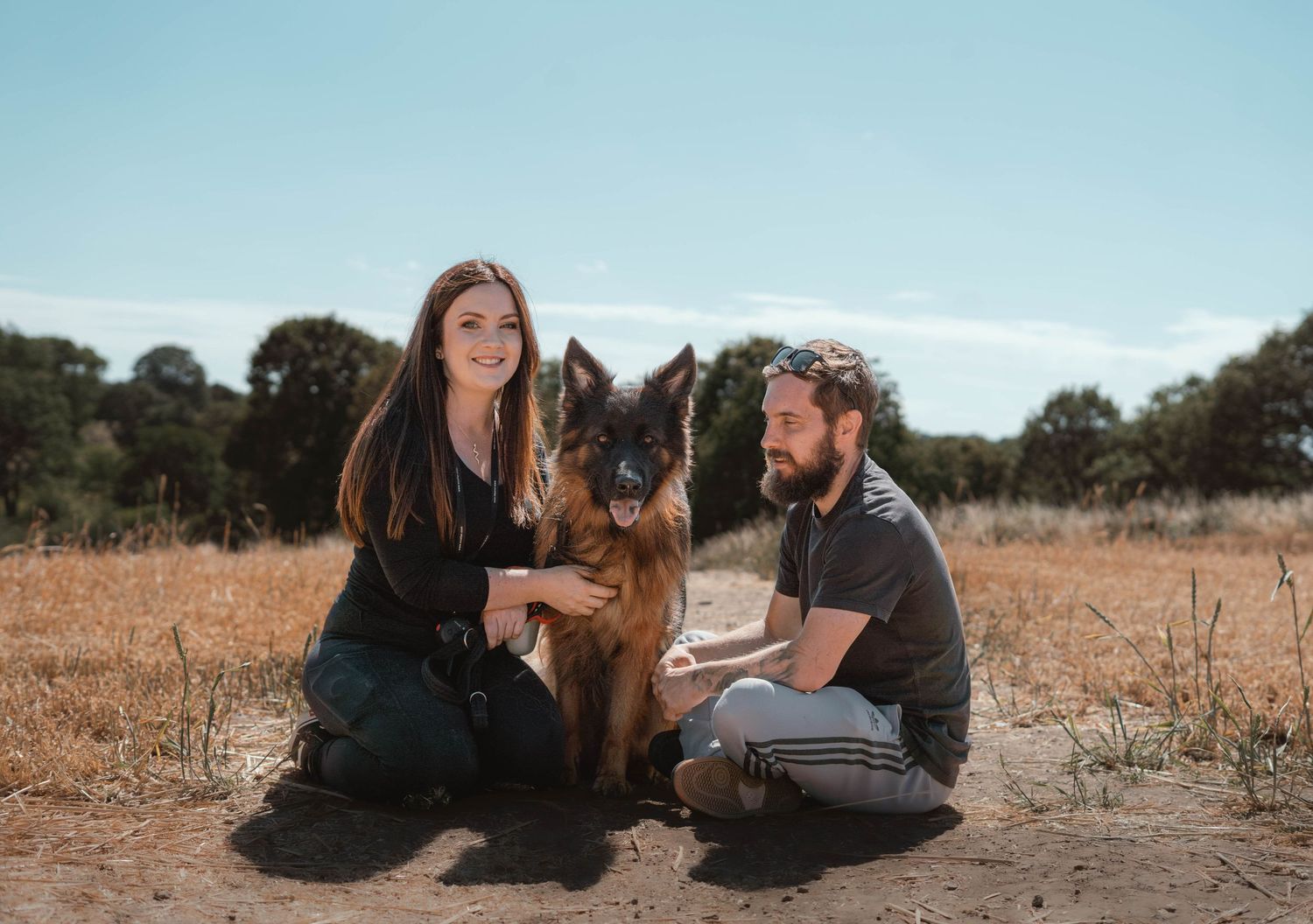 A couple kneels with their German Shepherd dog on a dirt path in a sunny field.