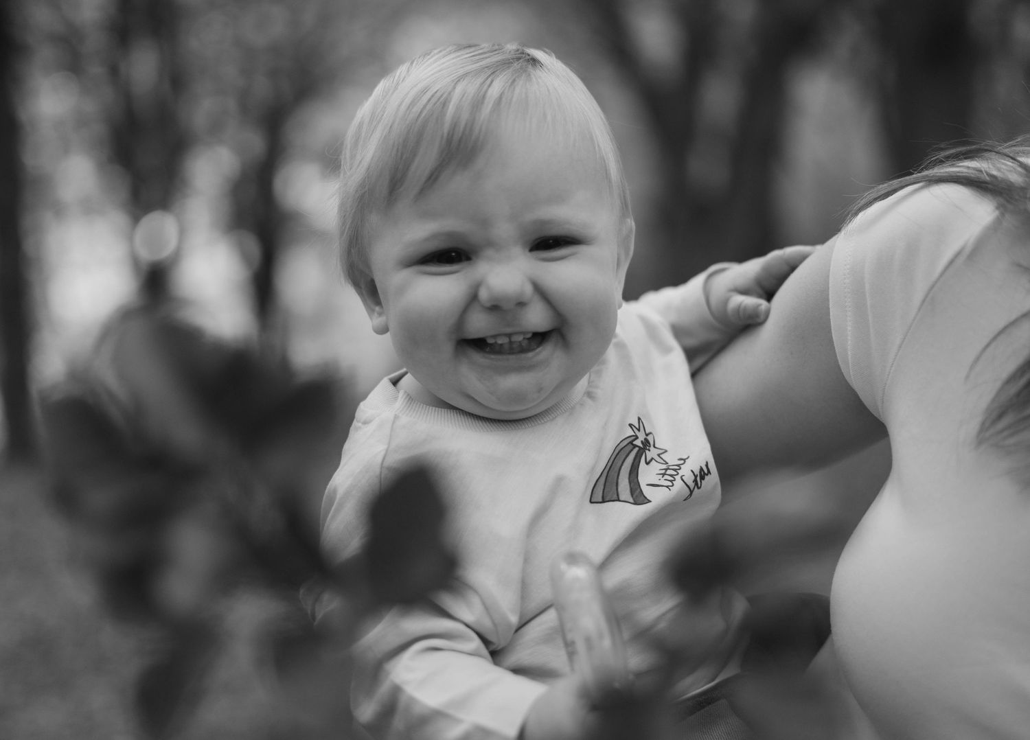 A joyful smiling baby being held up outdoors in a black and white portrait photo.