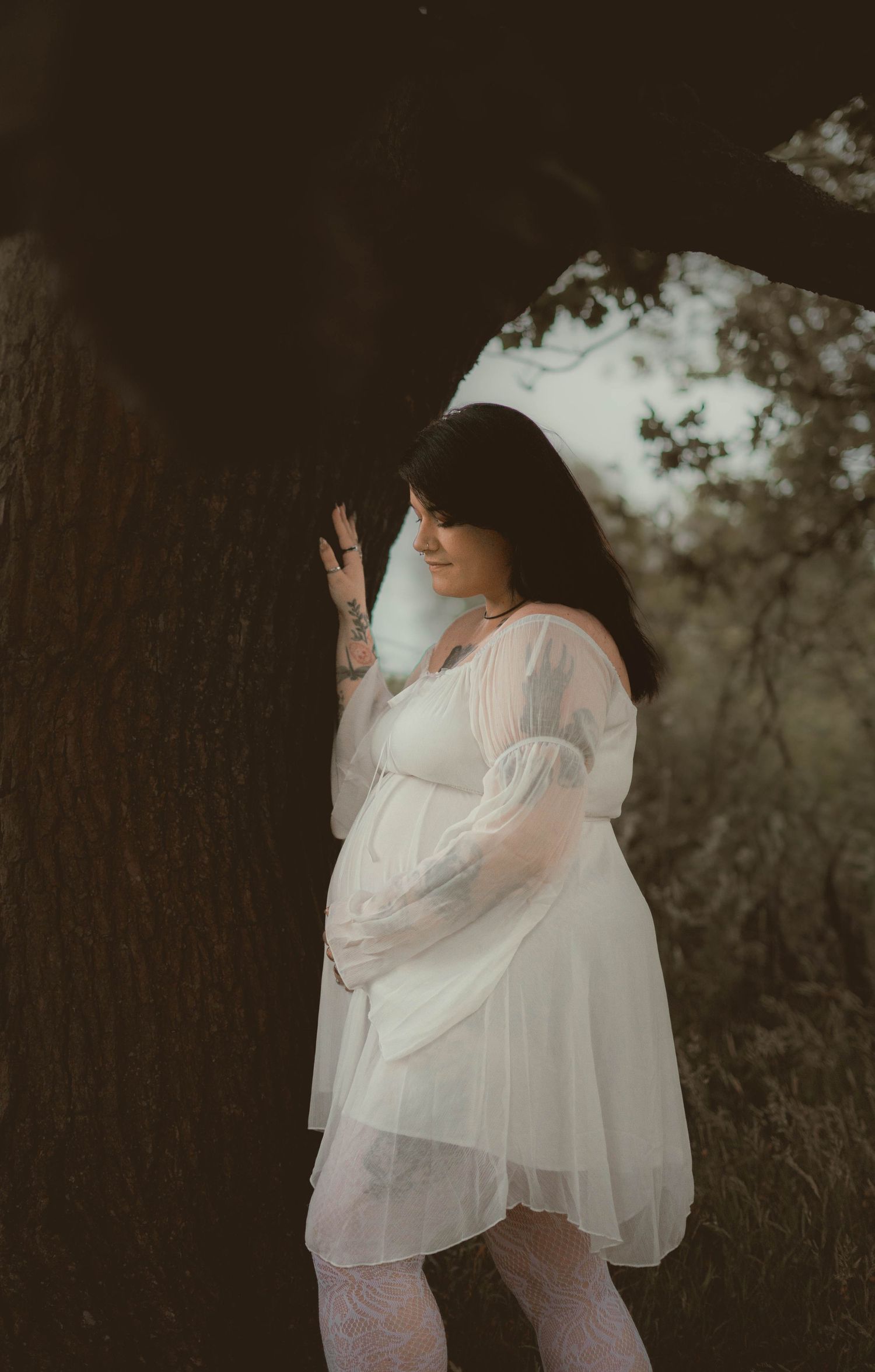 Pregnant woman in sheer white dress leaning against tree in dark forest setting.