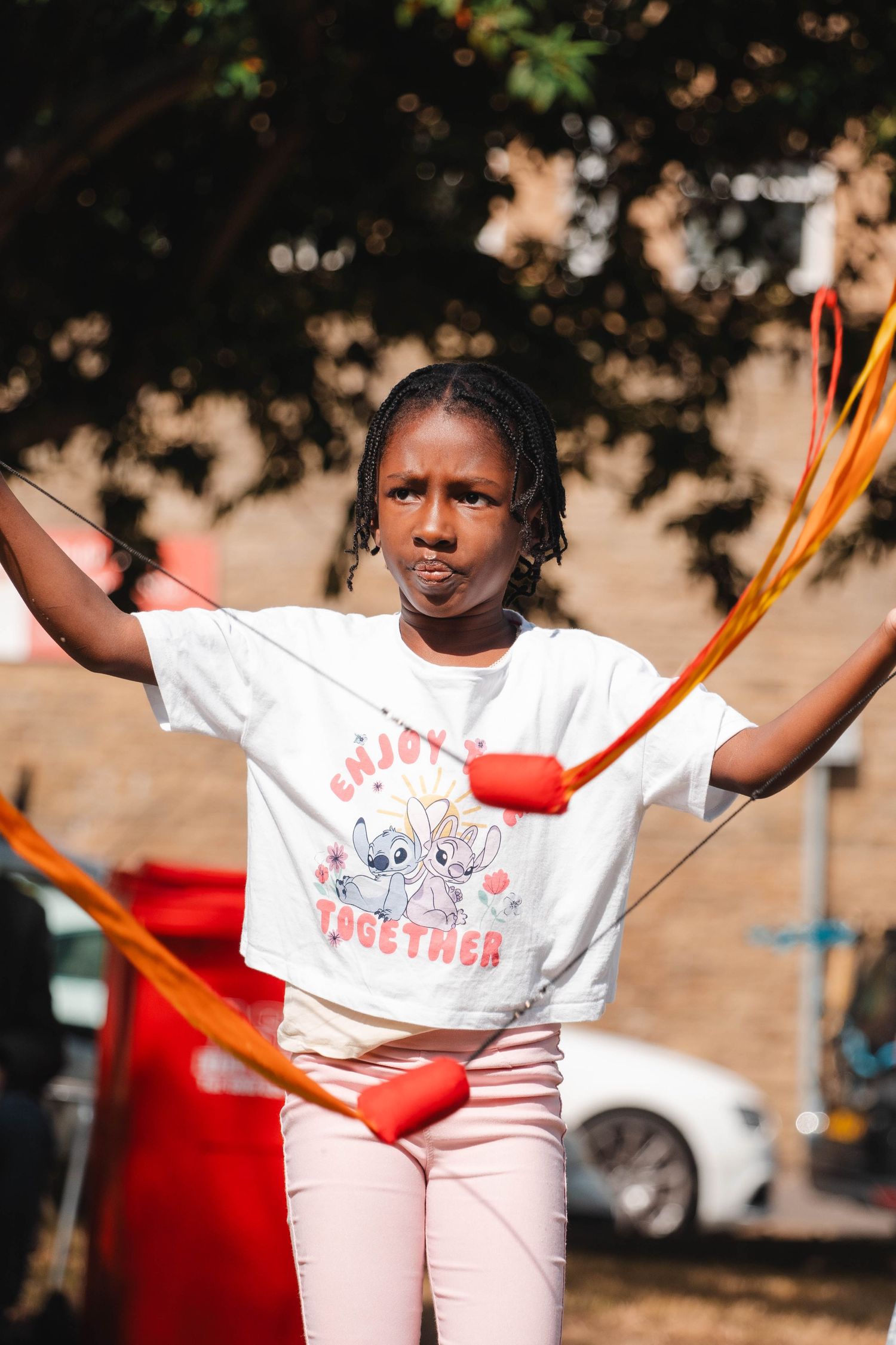 A girl in a white t-shirt holds orange jump rope handles outdoors in sunlight.