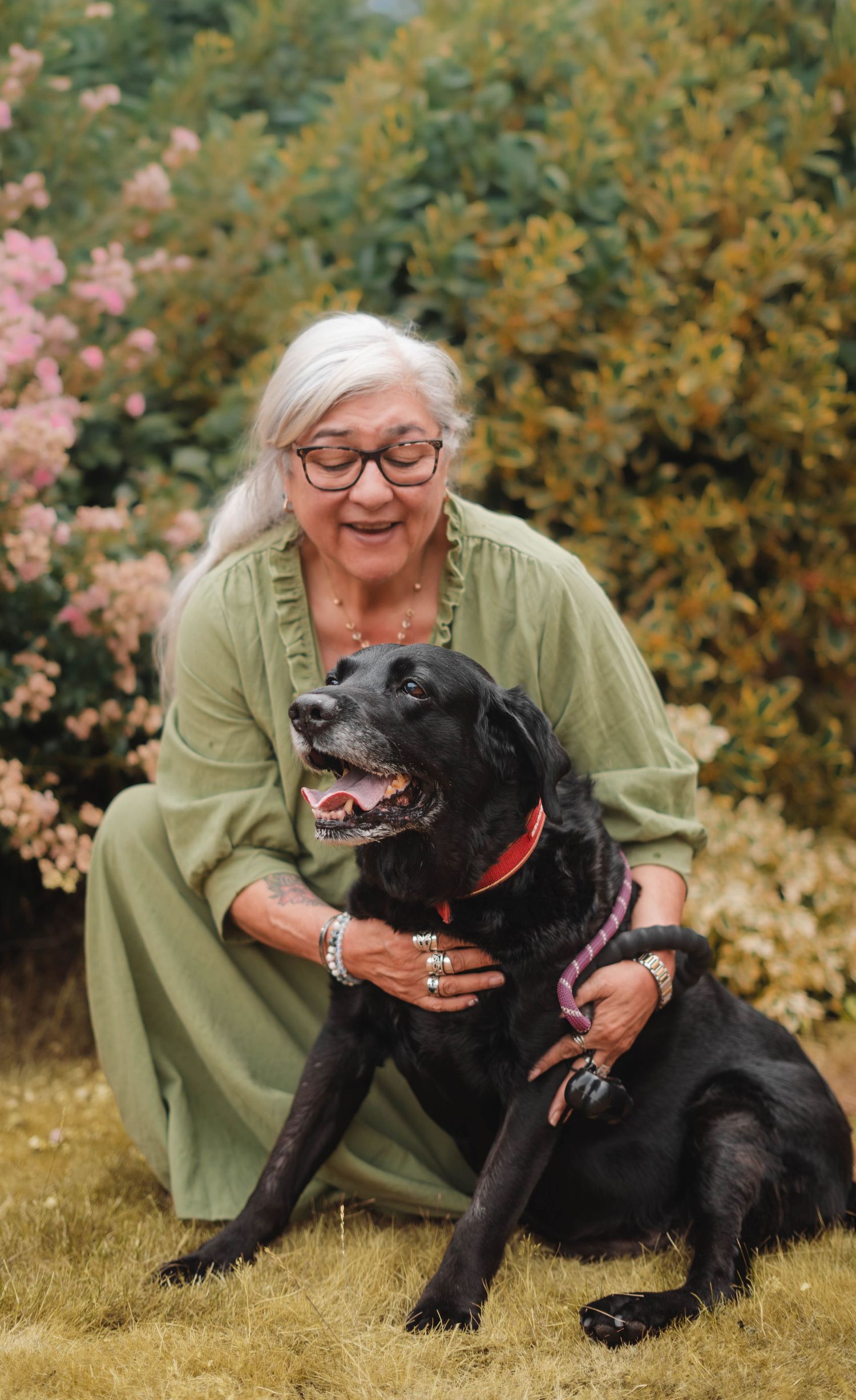 A smiling elderly woman with white hair cuddles a black Labrador dog in a colorful garden.