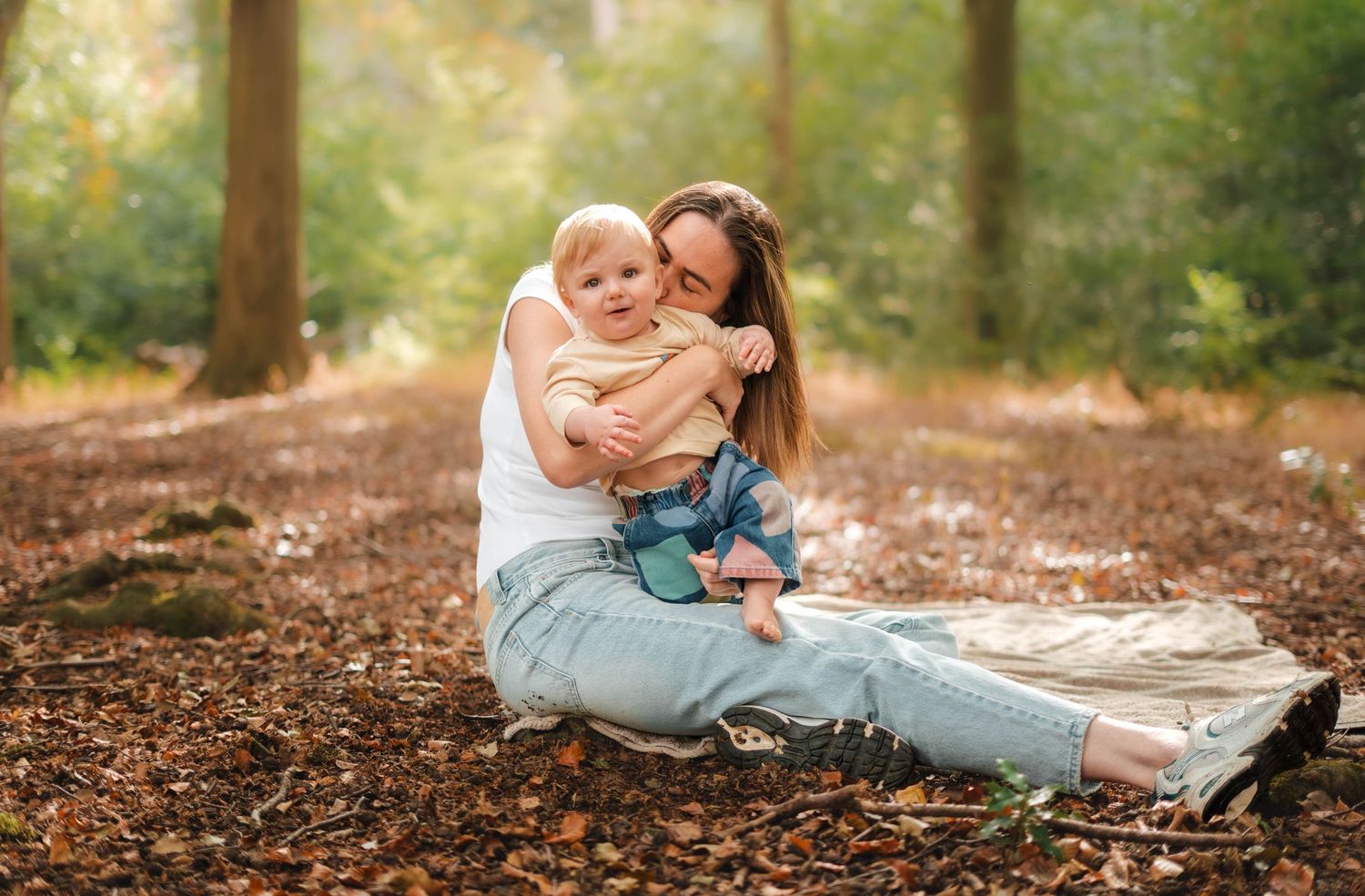 A loving mother sits on fallen autumn leaves hugging her toddler in a sunlit forest.