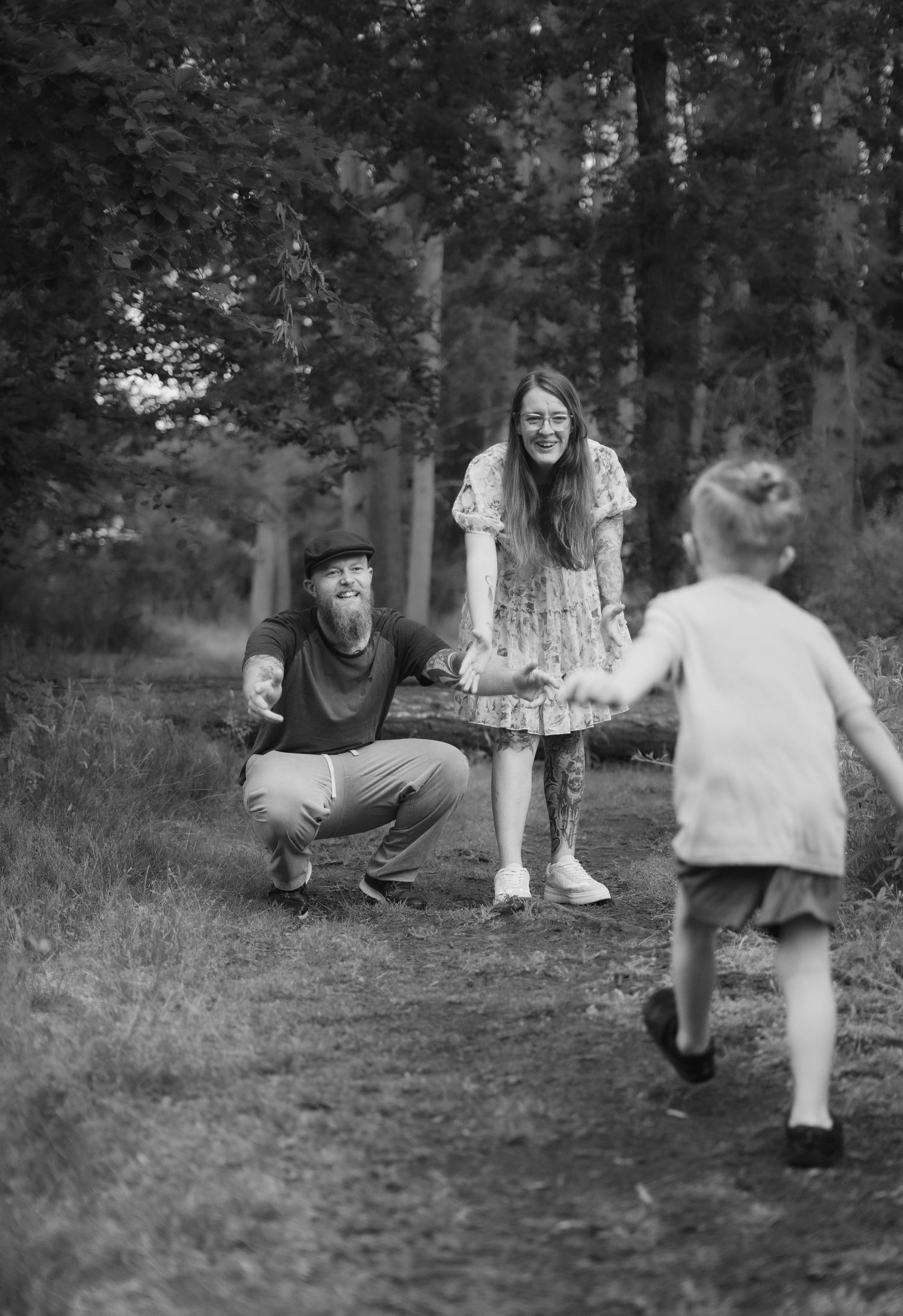 A black and white photo of parents crouching down as their young child runs toward them in a forest.
