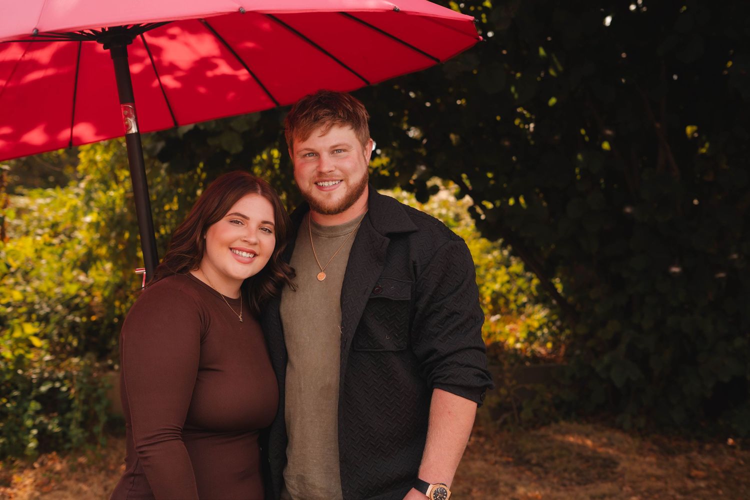 A smiling couple poses under a red umbrella surrounded by lush green foliage.