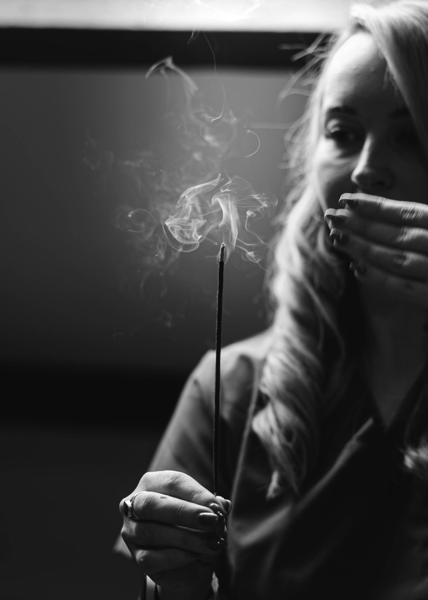 Black and white photo of a blonde woman holding a smoking incense stick in a dark room.