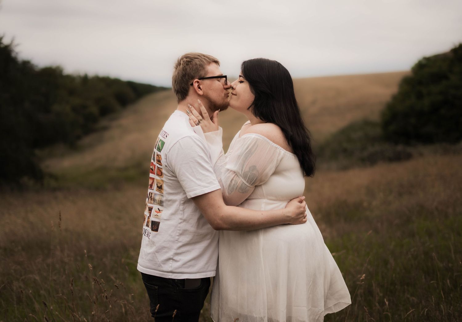 A couple shares a romantic kiss in an open field, the woman wearing a flowing white dress.