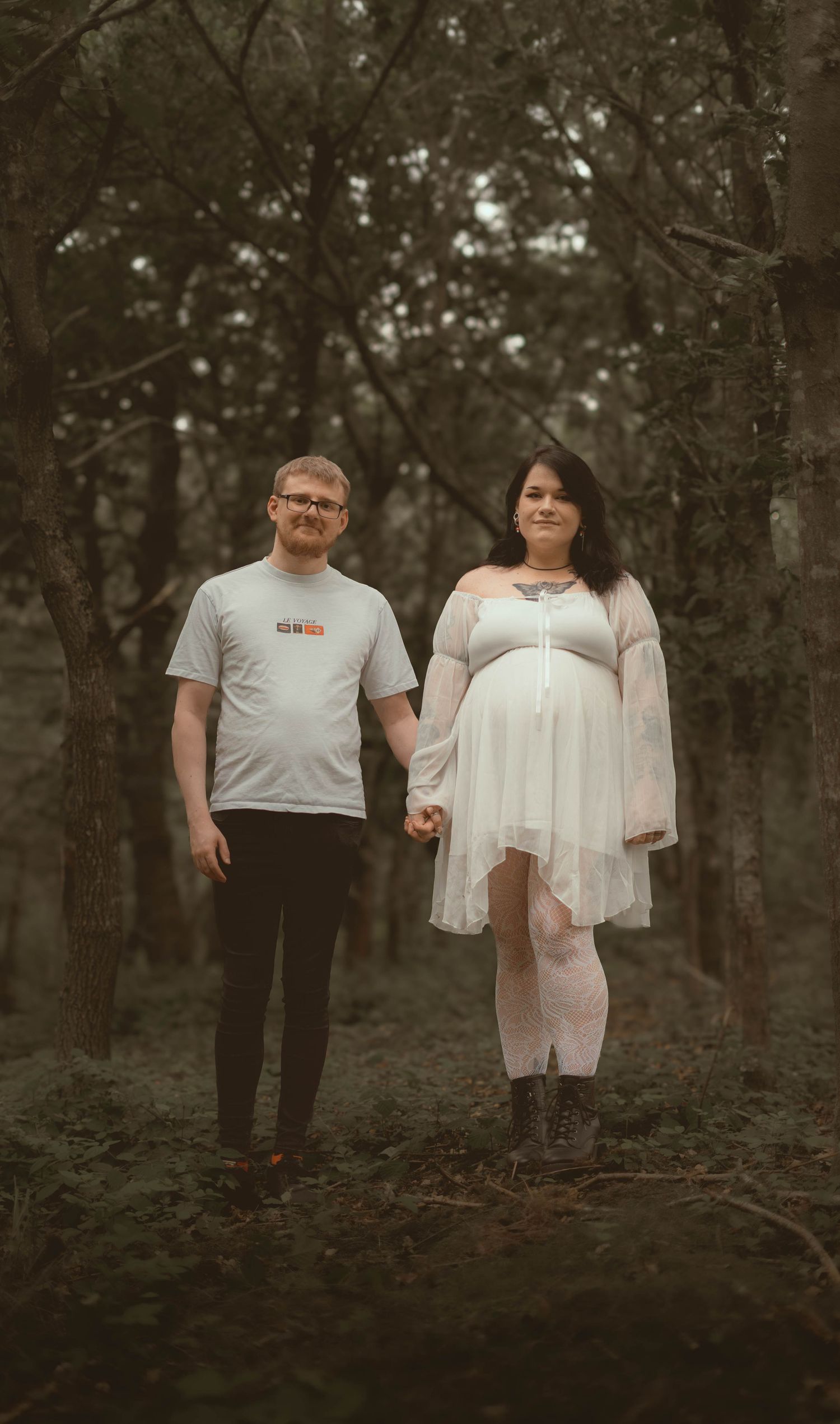 Expecting couple holding hands in misty forest, woman in white maternity dress and man in graphic tee.