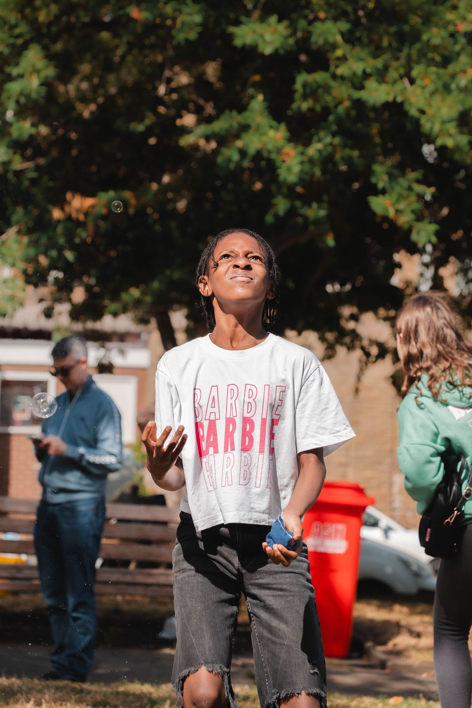 A young girl in a Barbie t-shirt looks up at bubbles floating in a sunny park.