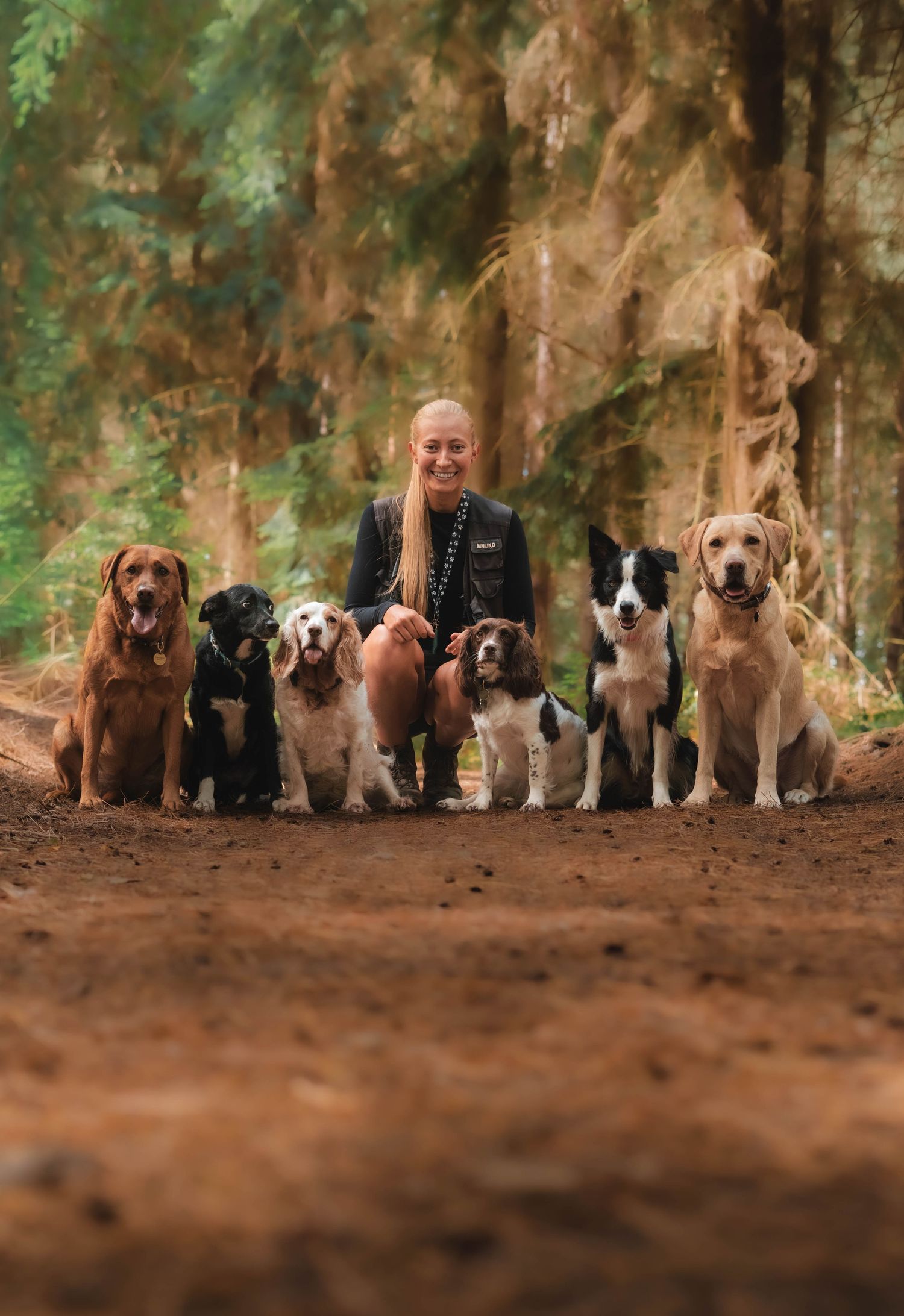 A smiling blonde woman poses with six dogs of various breeds on a forest dirt path surrounded by tall trees.