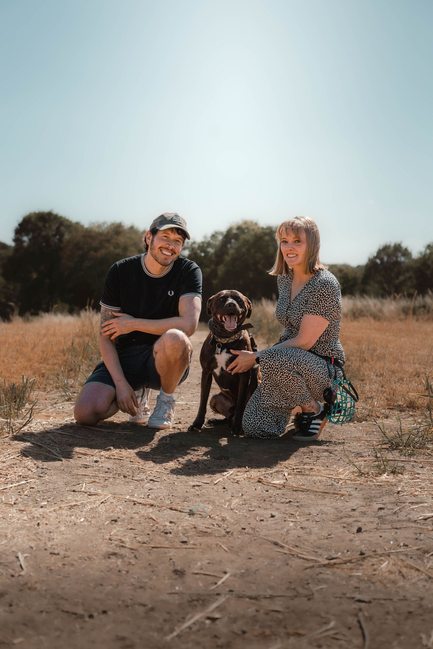 A smiling couple kneels beside their chocolate Labrador dog on a sunny dry dirt path in an open field.