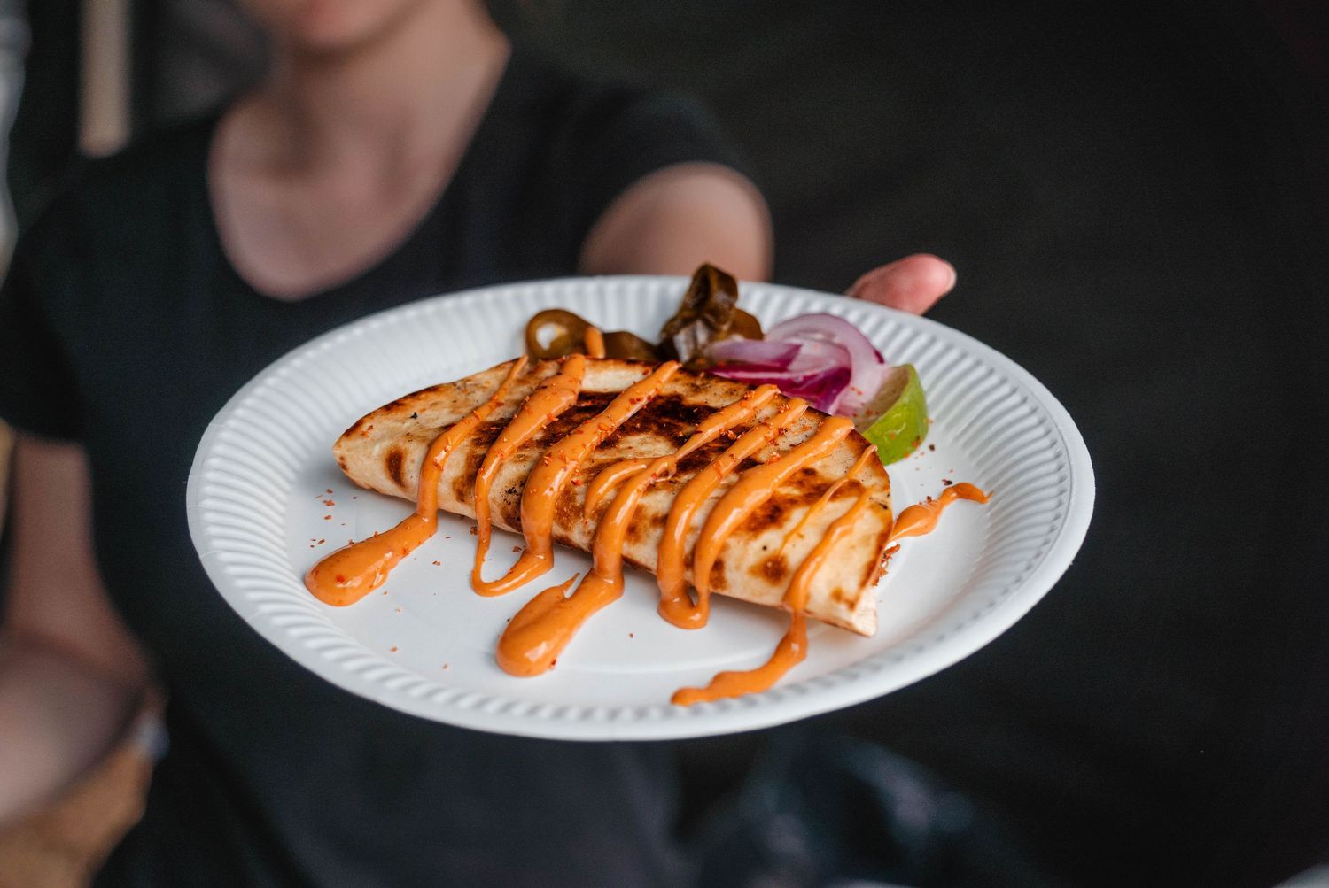 Person presenting grilled halloumi with sriracha drizzle and garnish on white plate.