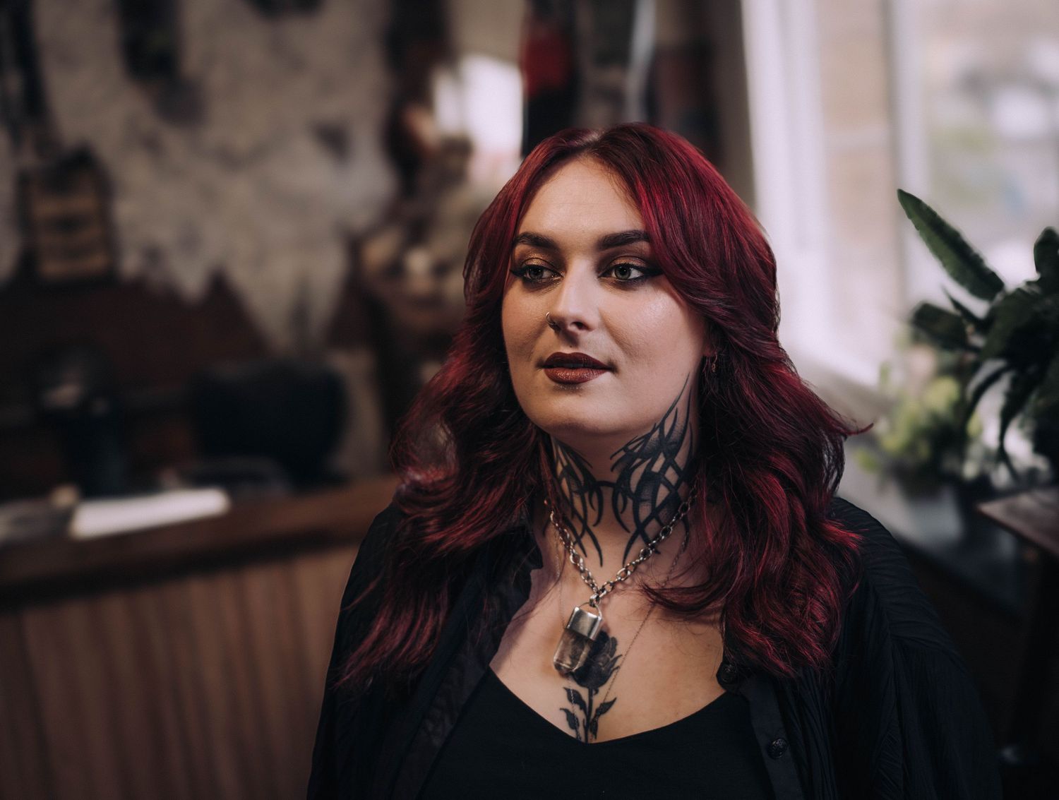 Close-up portrait of a tattooed woman with red hair and neck tattoos inside a tattoo studio.