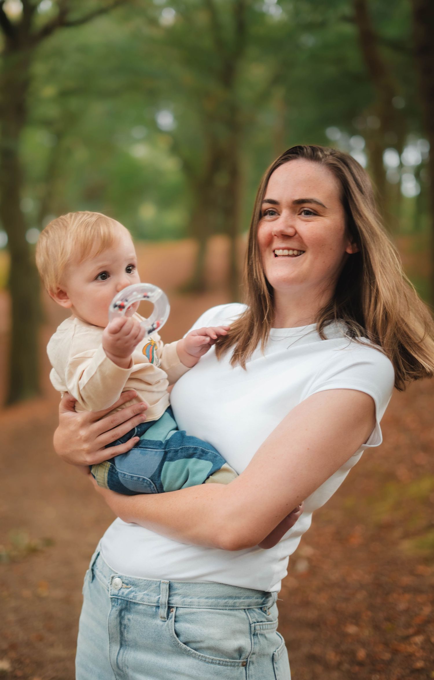 Smiling young mother holding baby with pacifier on autumn forest path.