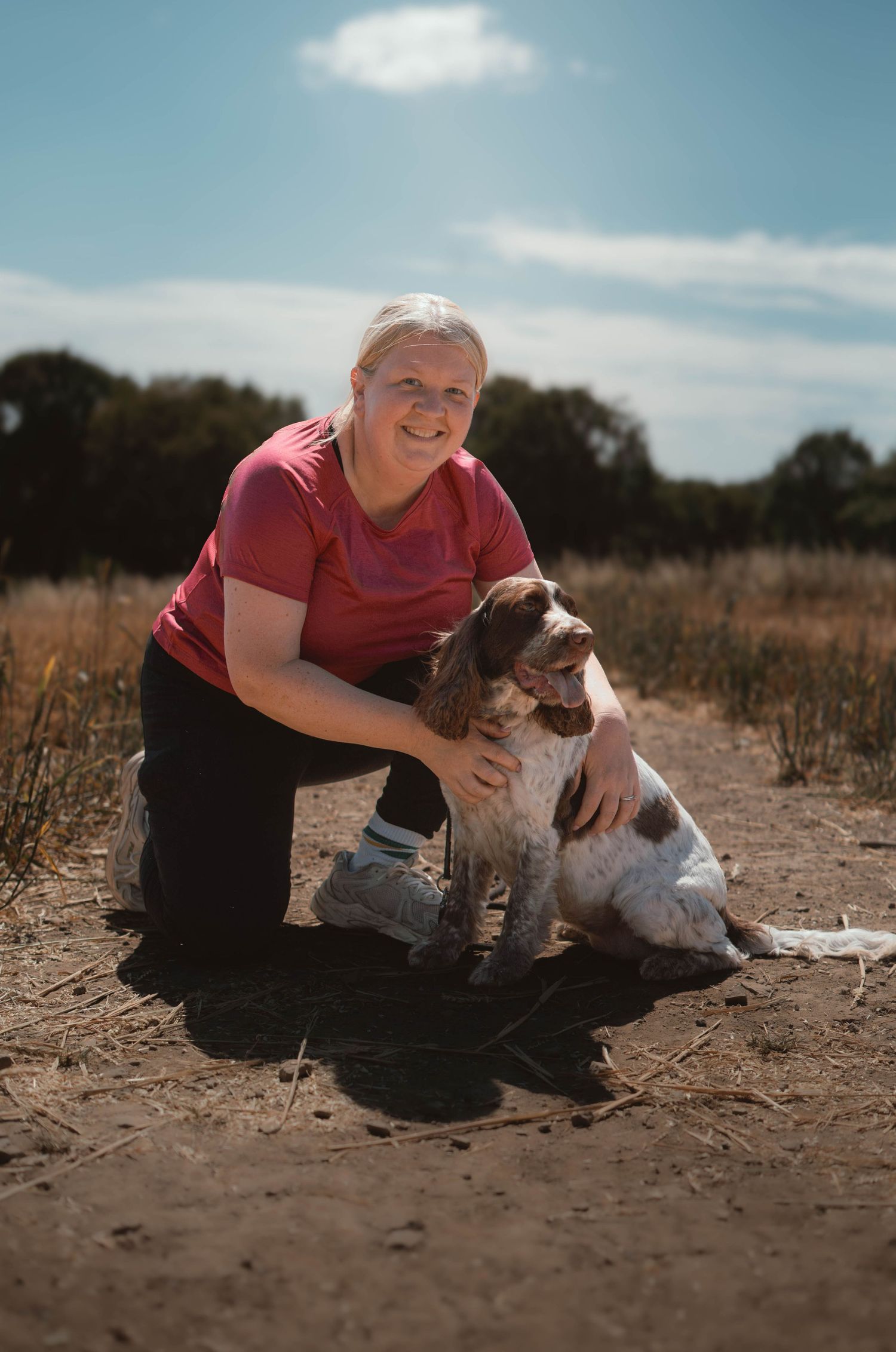 Smiling blonde woman in pink shirt hugging a Springer Spaniel dog on a sunny dirt path.