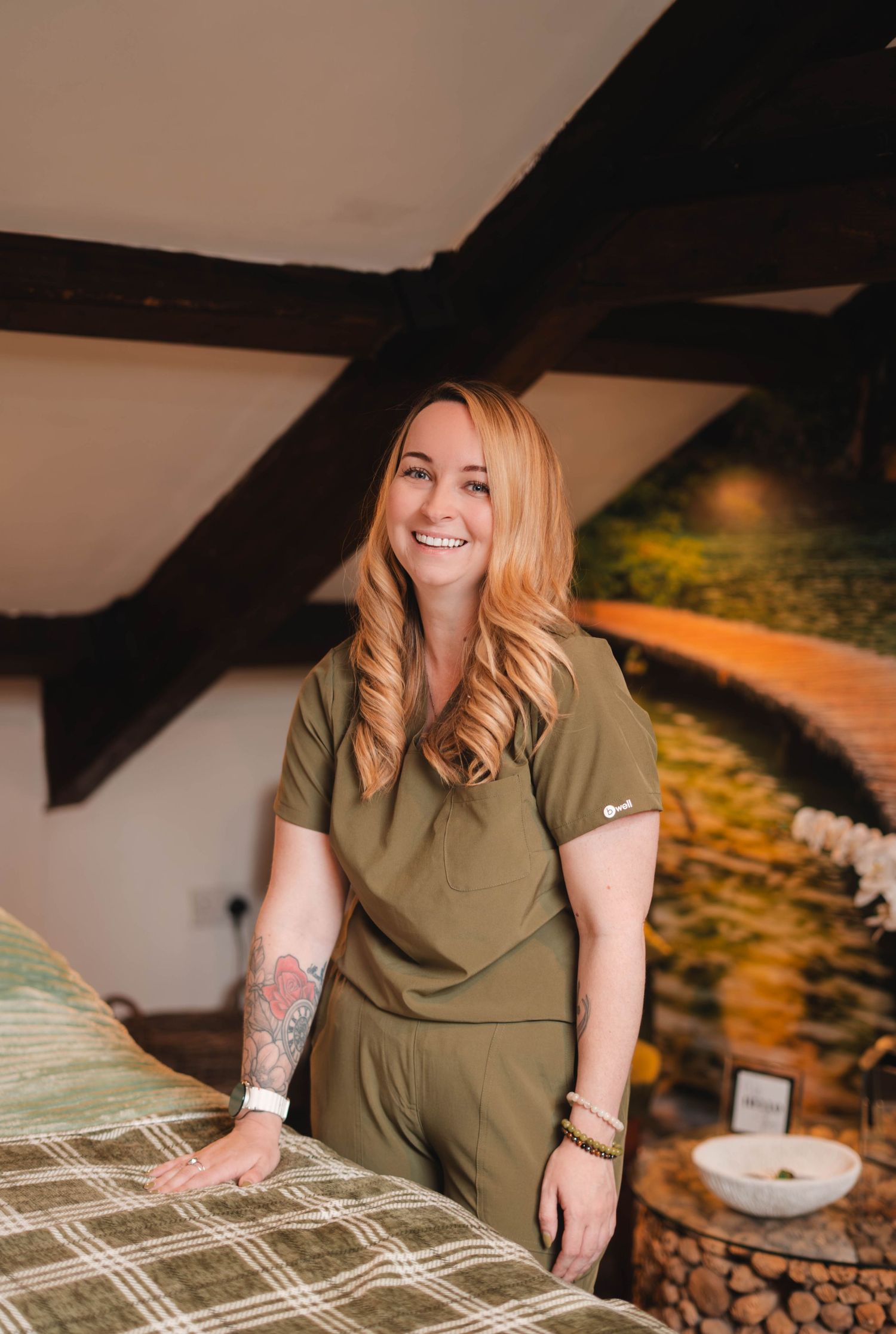 Smiling woman in olive green scrubs stands beside a massage table in a cozy spa with wooden beam ceiling.