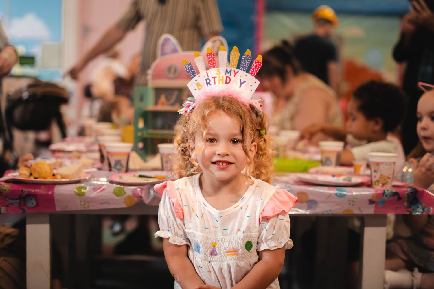 Young curly-haired girl wearing birthday crown sitting at decorated party table.