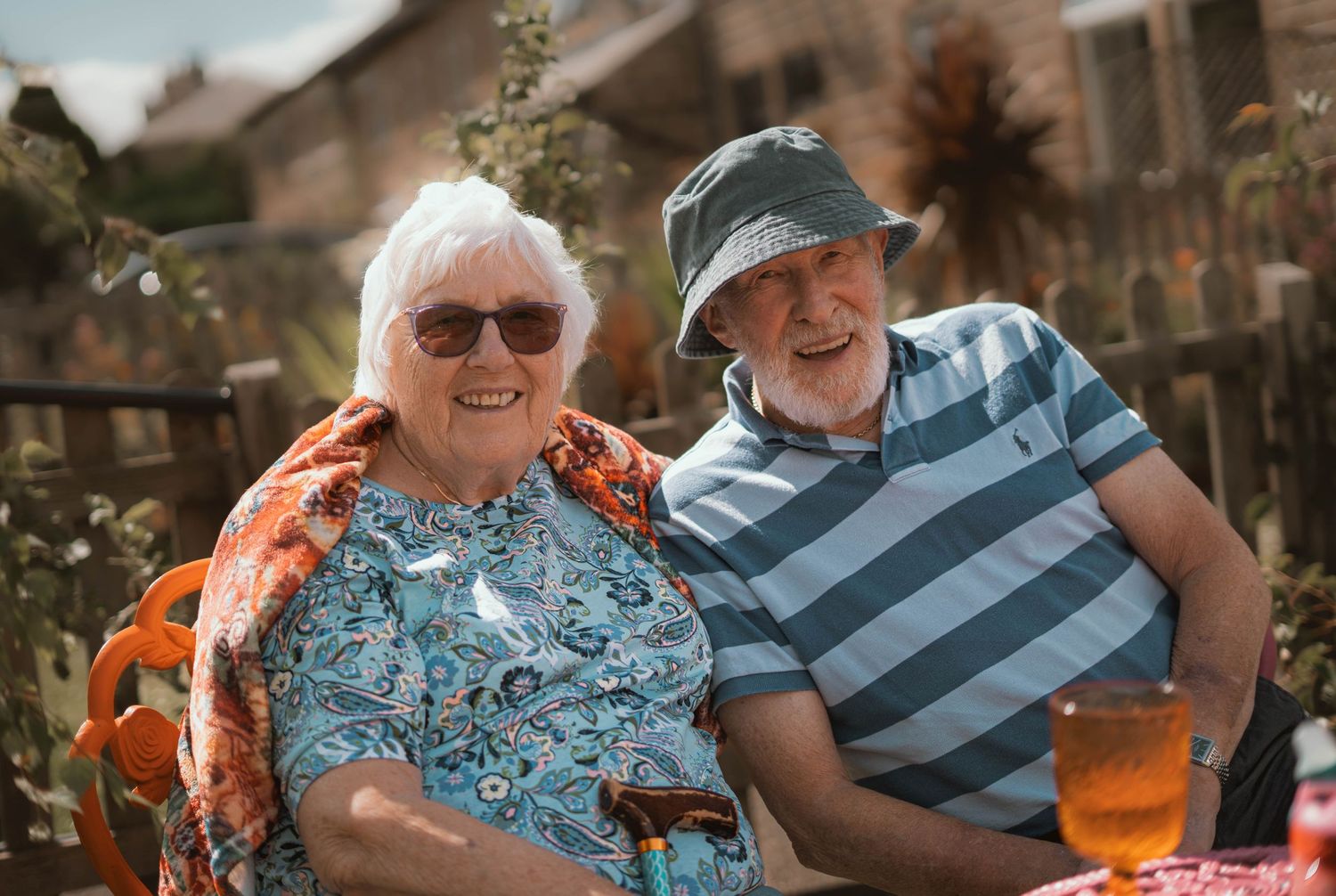 Elderly couple smiling outdoors in sunny garden, woman in floral top with sunglasses, man in striped shirt and bucket hat.