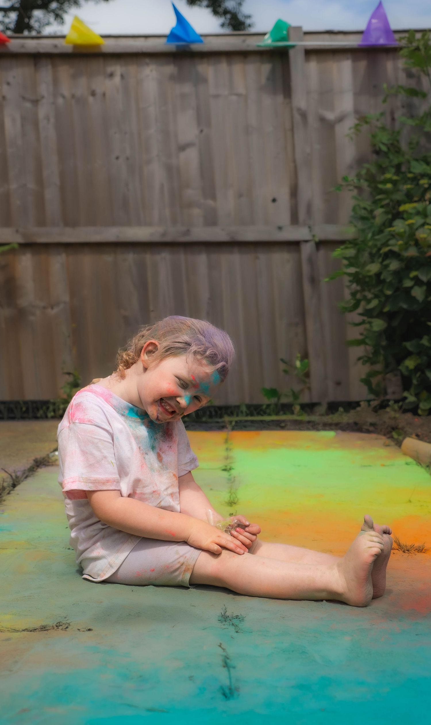 Happy child sitting on colorful powder-covered ground laughing during outdoor Holi color festival celebration.
