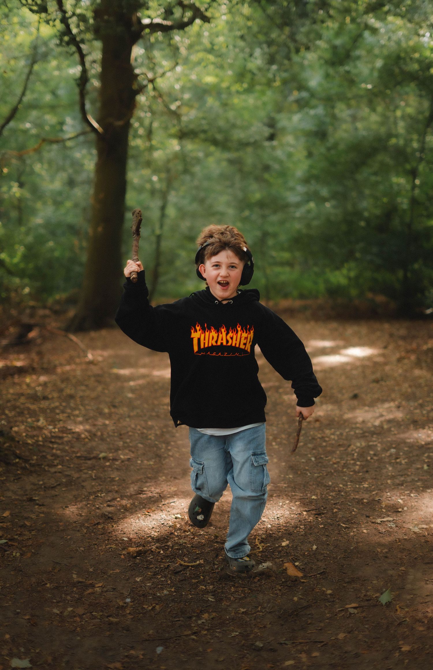 A joyful boy runs through a forest path wearing a black Thrasher hoodie and jeans, holding sticks with excitement.