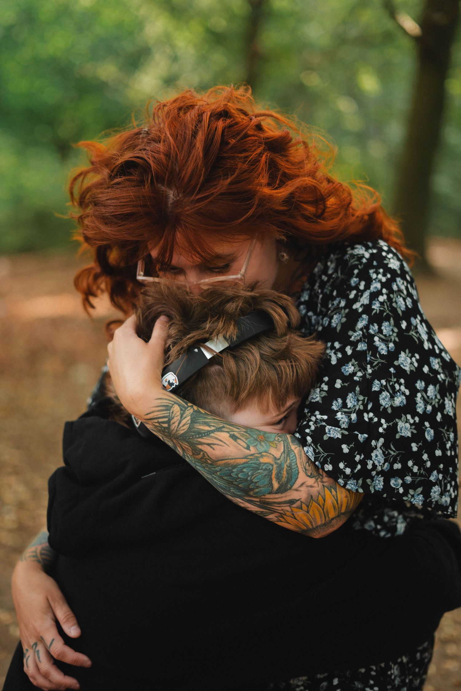Tattooed red-haired woman tenderly hugging a child in a woodland setting.