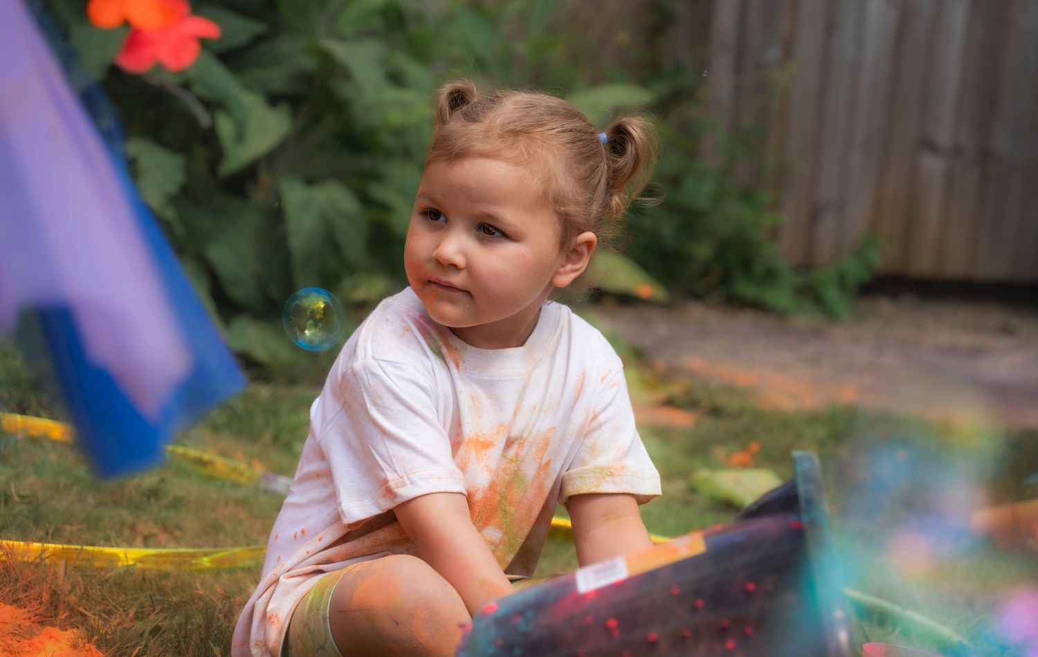 Toddler girl sitting on grass outdoors during a colorful Holi powder celebration in a garden.