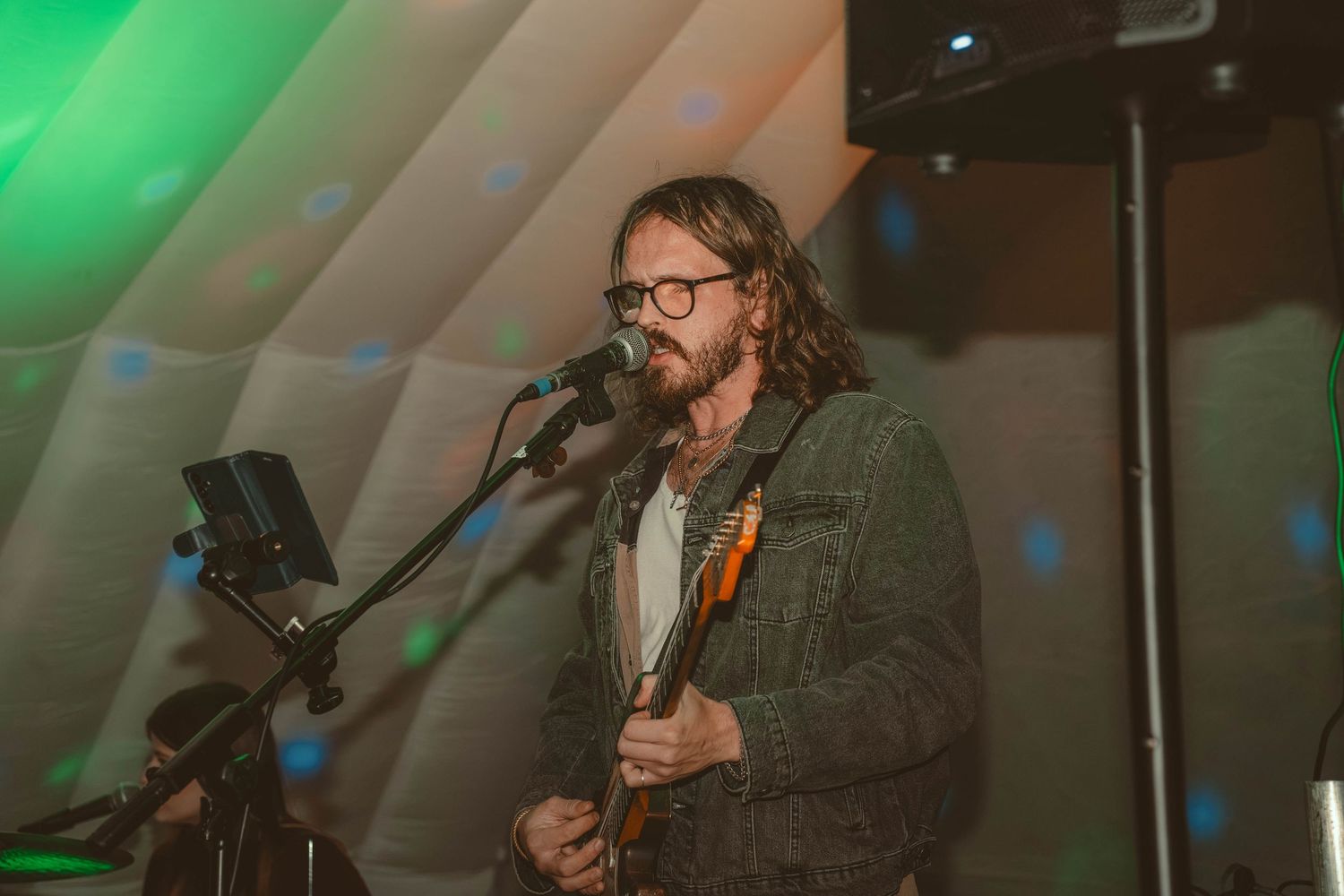 Long-haired male guitarist singing into microphone on stage with green spotlights.