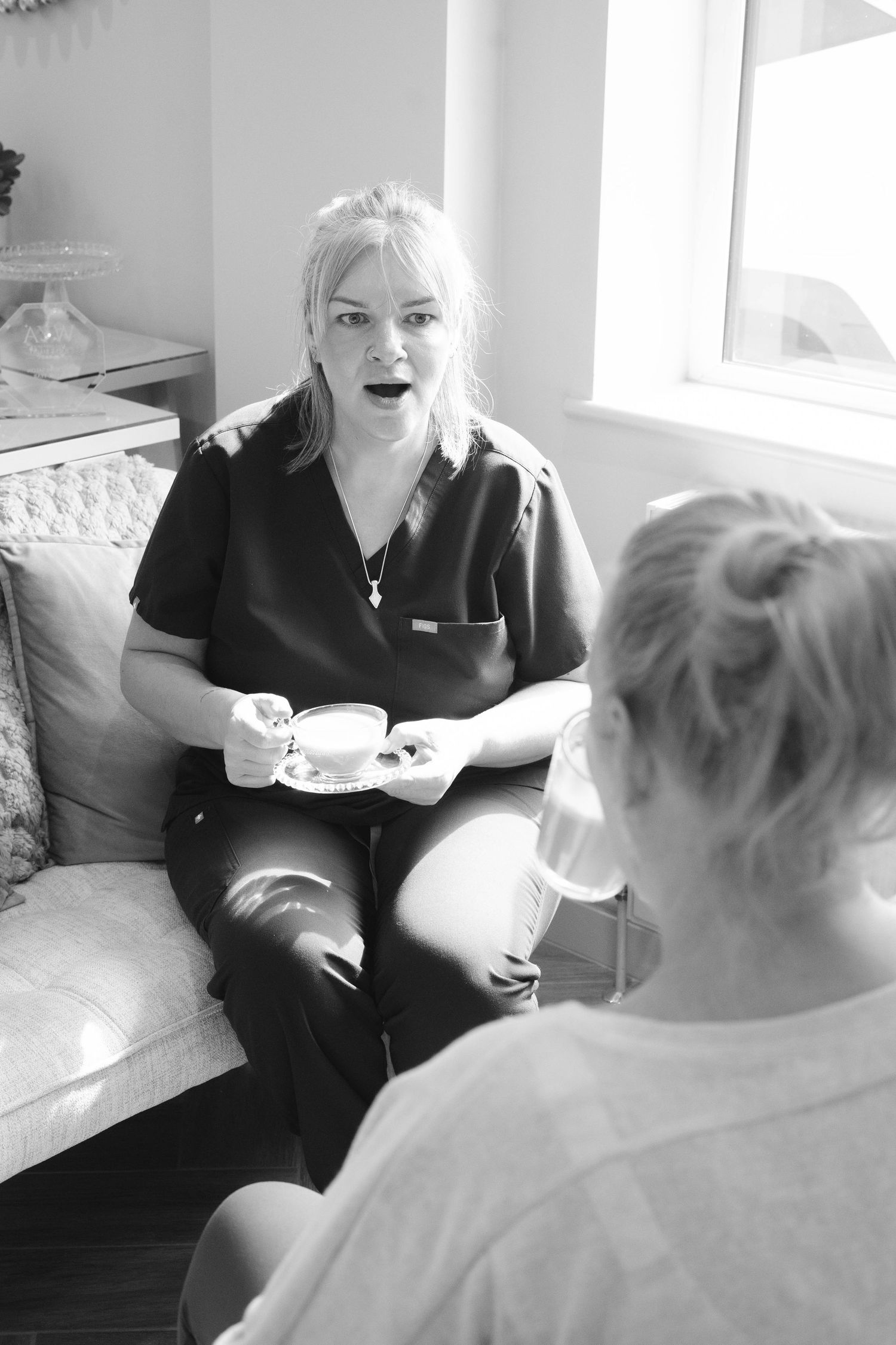 A blonde woman in black scrubs holds a teacup while consulting with a client on a bright sofa.