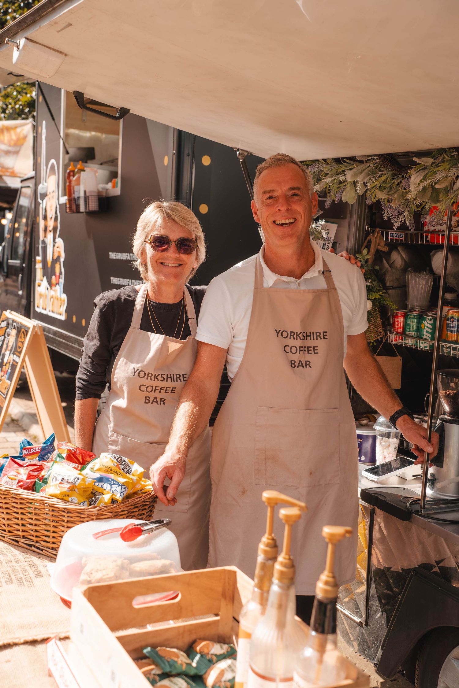 Two smiling staff in Yorkshire Coffee Bar aprons serving at outdoor market food stall.