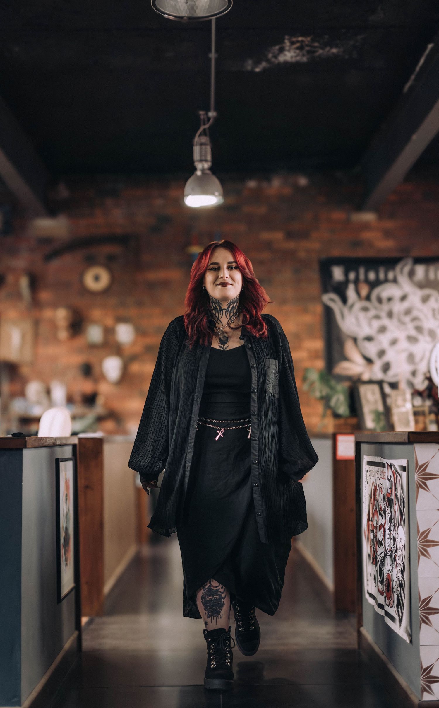 Heavily tattooed woman with red hair wearing all black walks through a rustic brick tattoo studio.