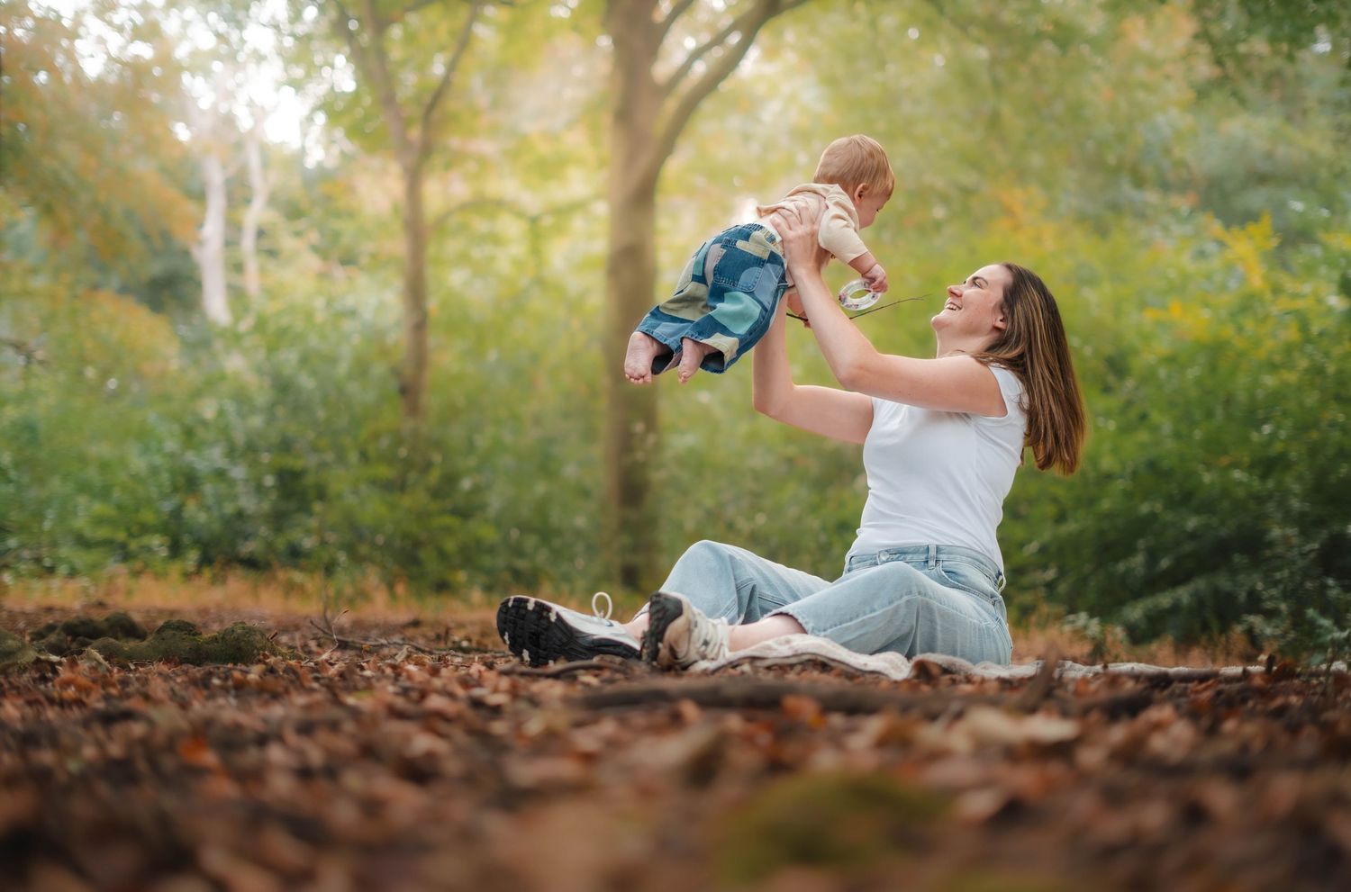 A young mother sits on autumn leaves in a park, joyfully lifting her baby up high.
