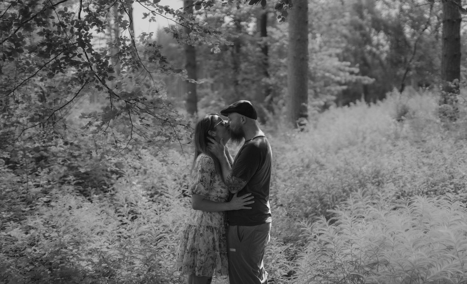 A couple shares a romantic kiss surrounded by lush green forest in a black and white photo.