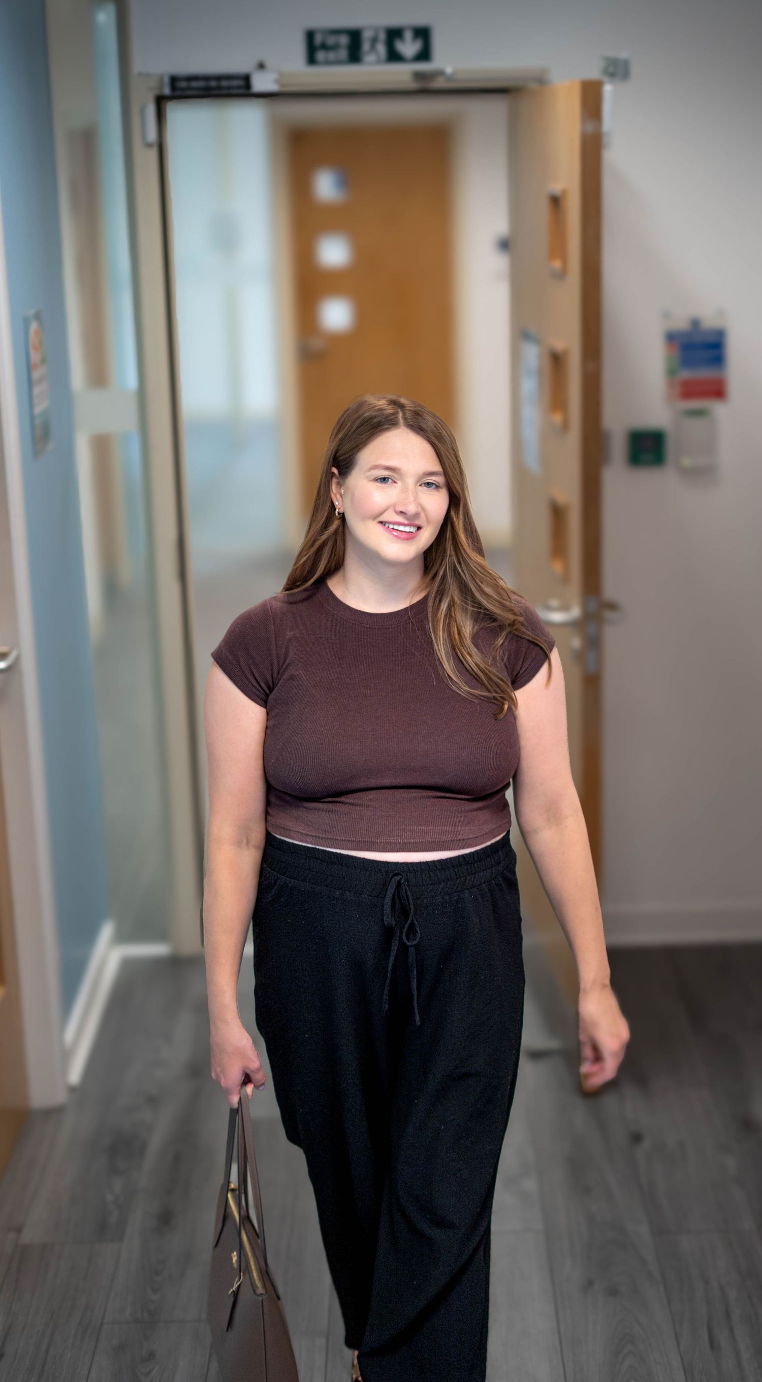 Young woman in brown crop top and black trousers walking confidently through modern office corridor.
