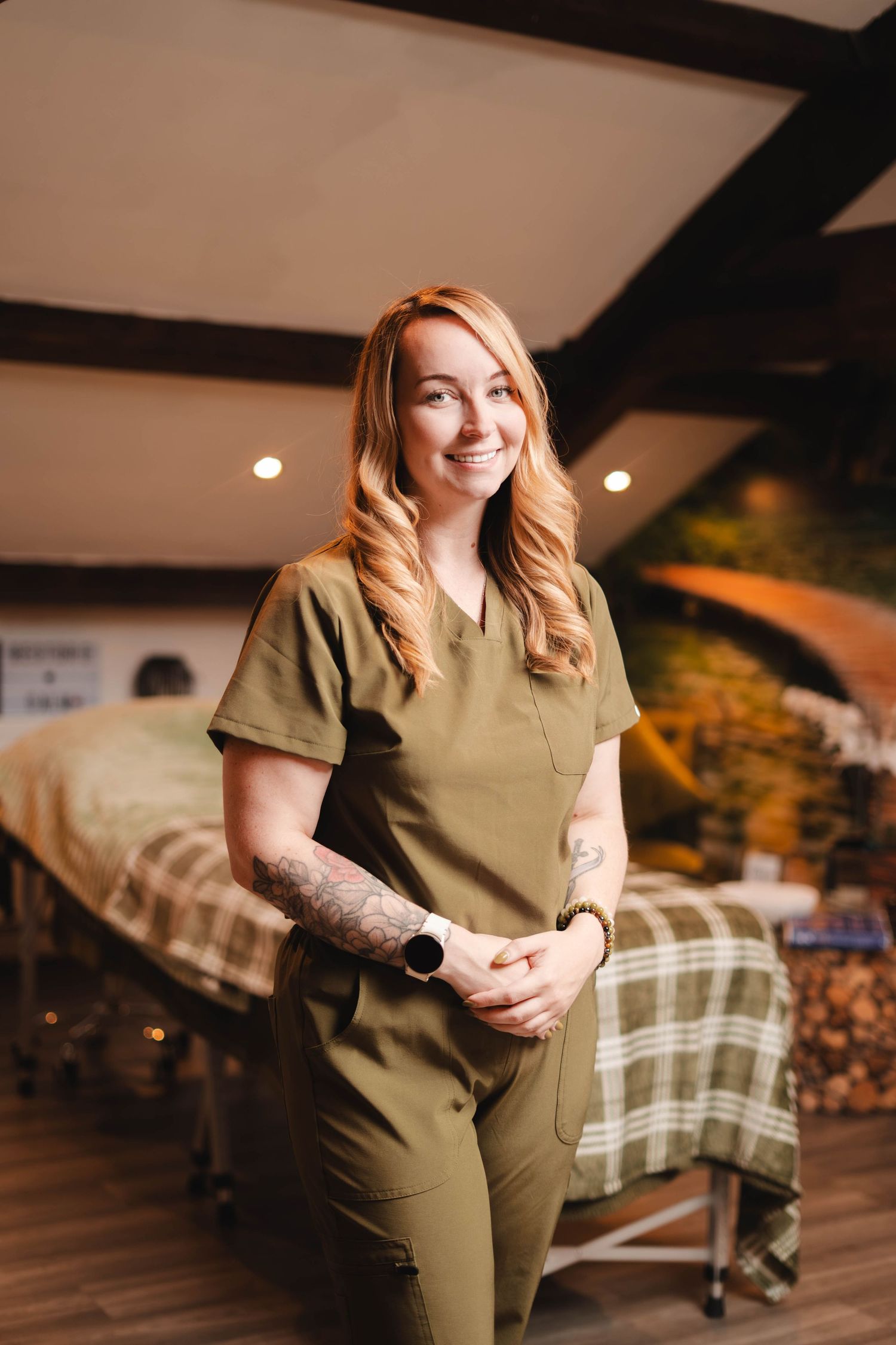Smiling female massage therapist in olive green scrubs with tattoos standing in a spa treatment room.