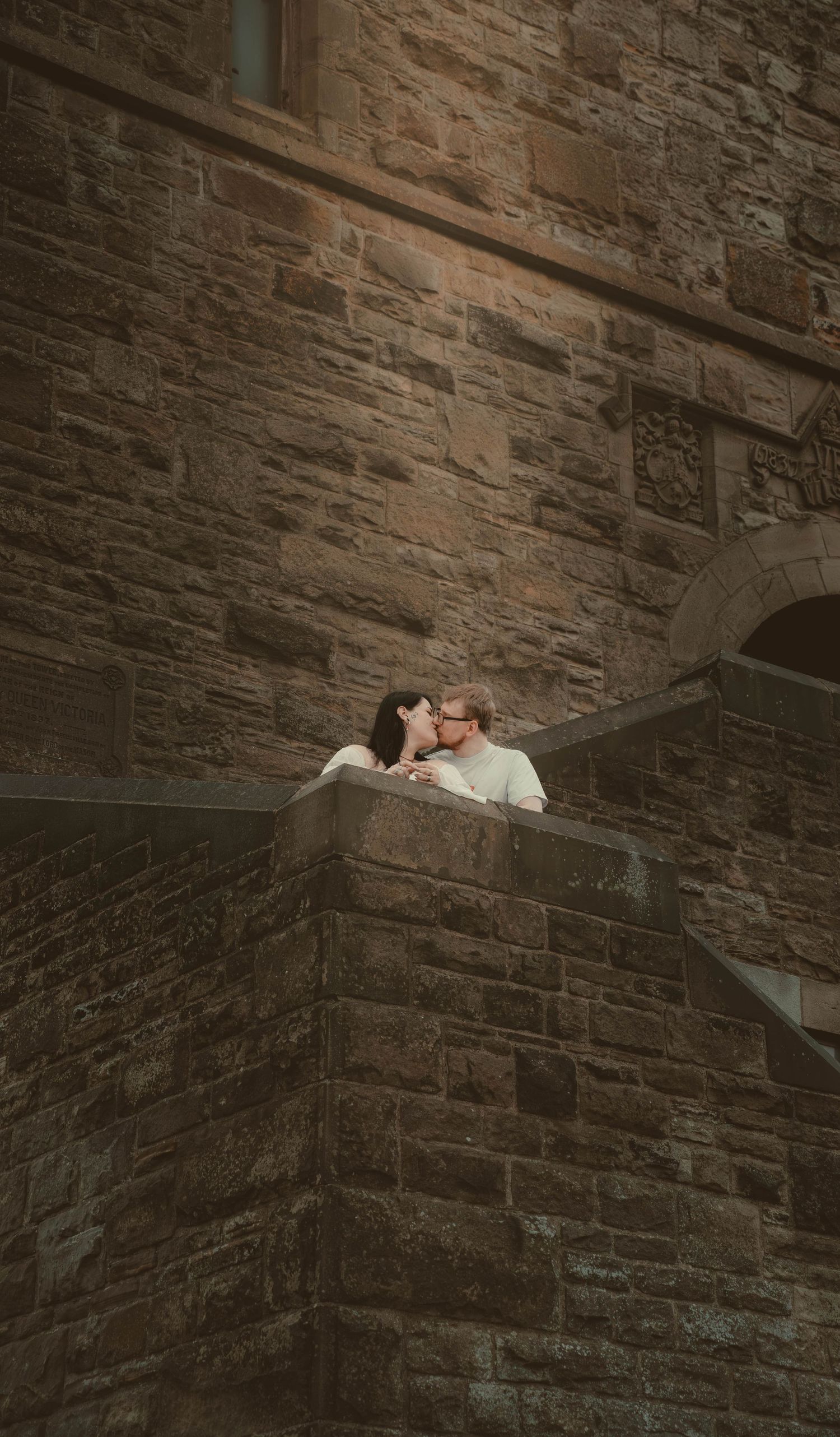 Couple sharing a kiss on stone castle balcony with rustic brick wall backdrop.