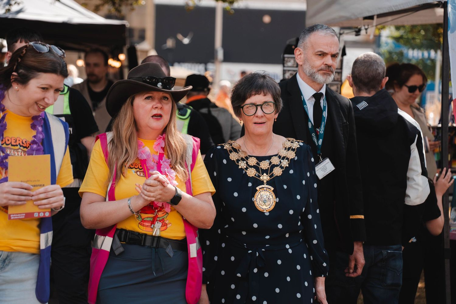 A group of people including a woman wearing a mayoral chain walk through a festival.