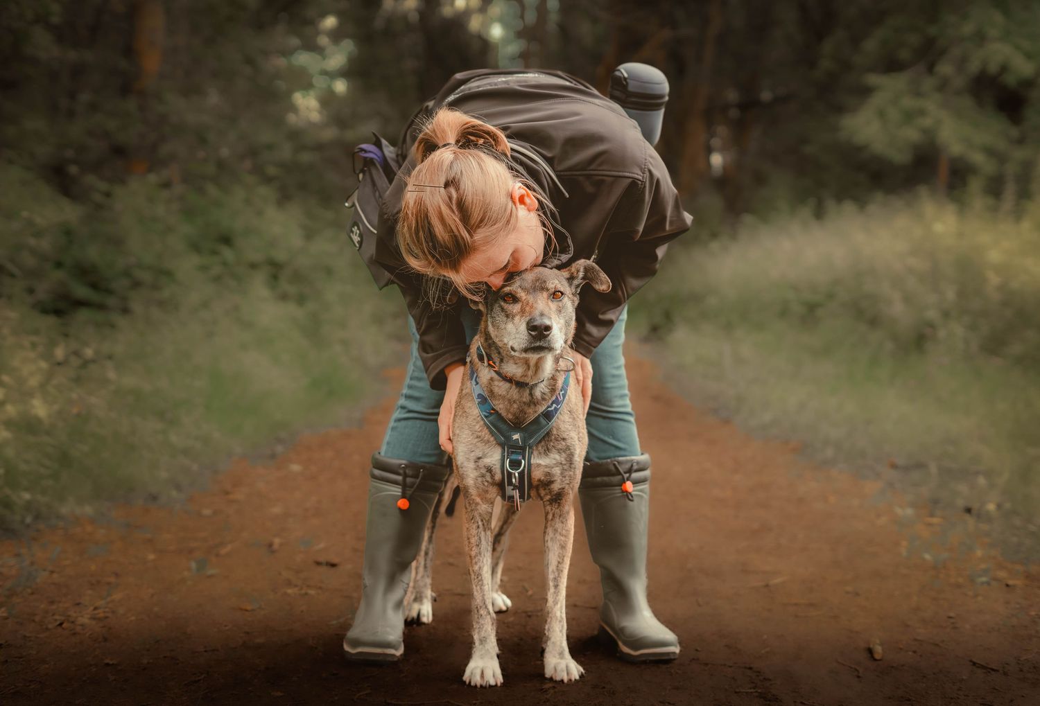 Woman bending down to hug her small dog on a woodland trail path.