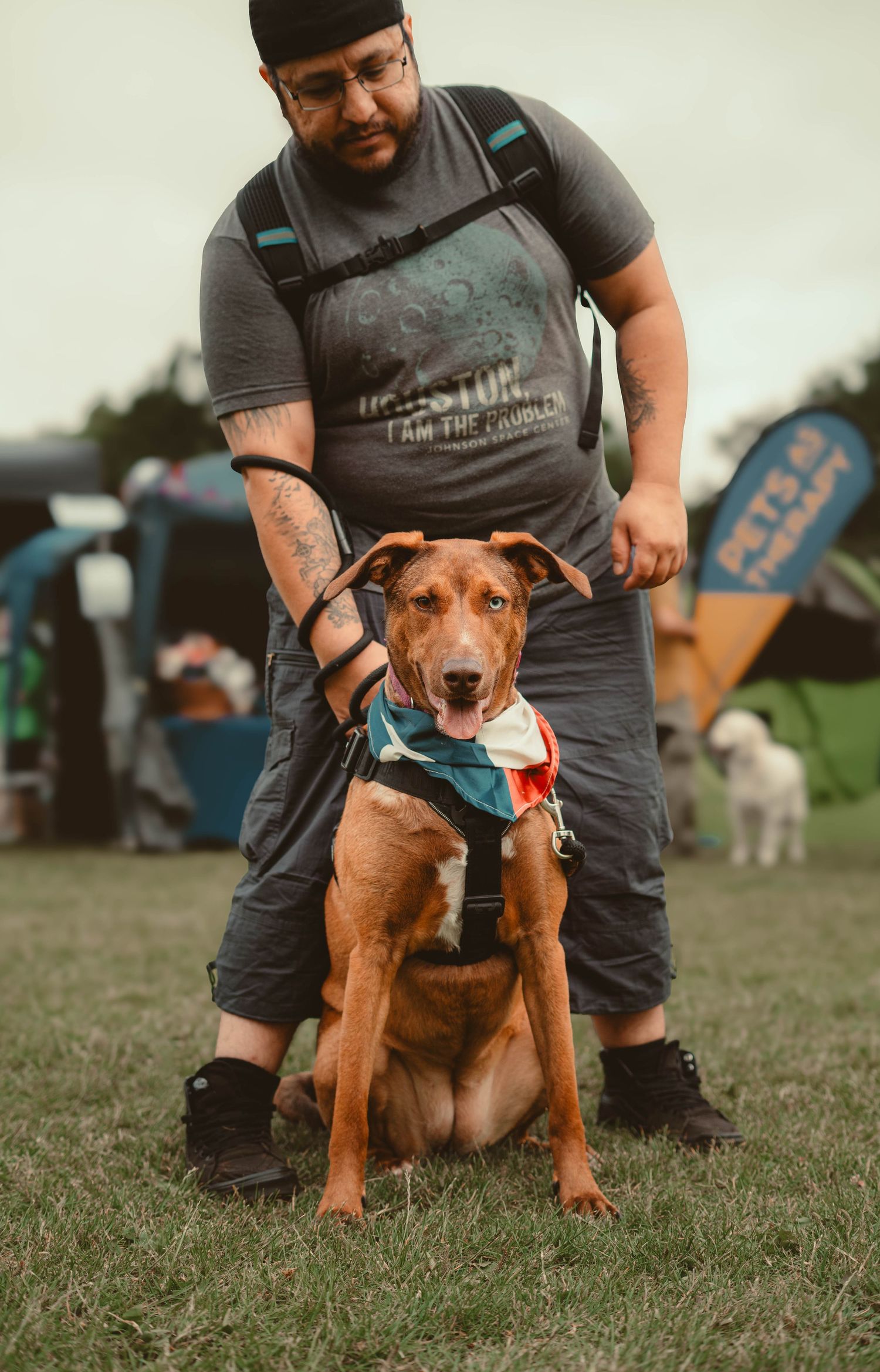 Man with backpack standing behind brown Pitbull mix dog wearing bandana on leash at outdoor festival.